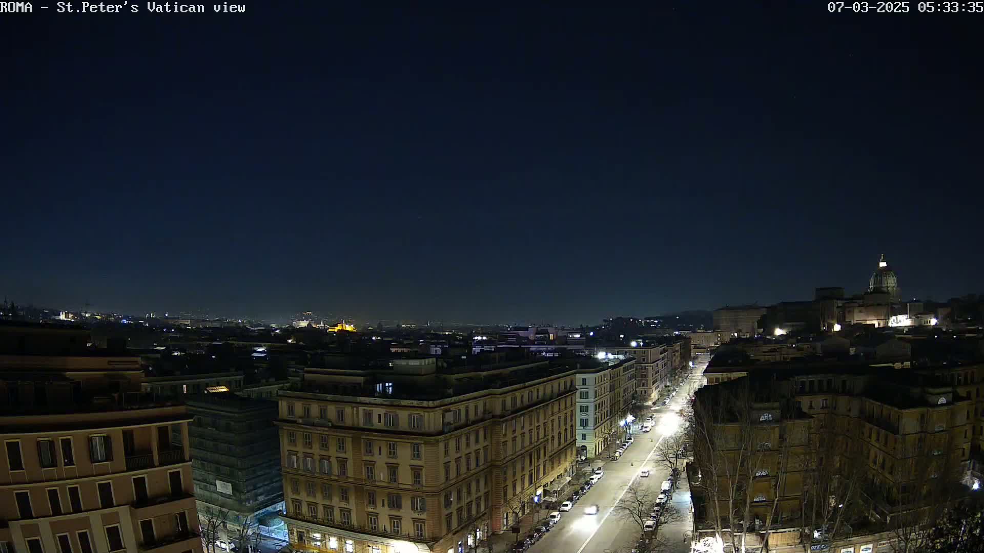 A nighttime aerial view of a city, showing a street with cars and buildings illuminated under a clear, dark sky.