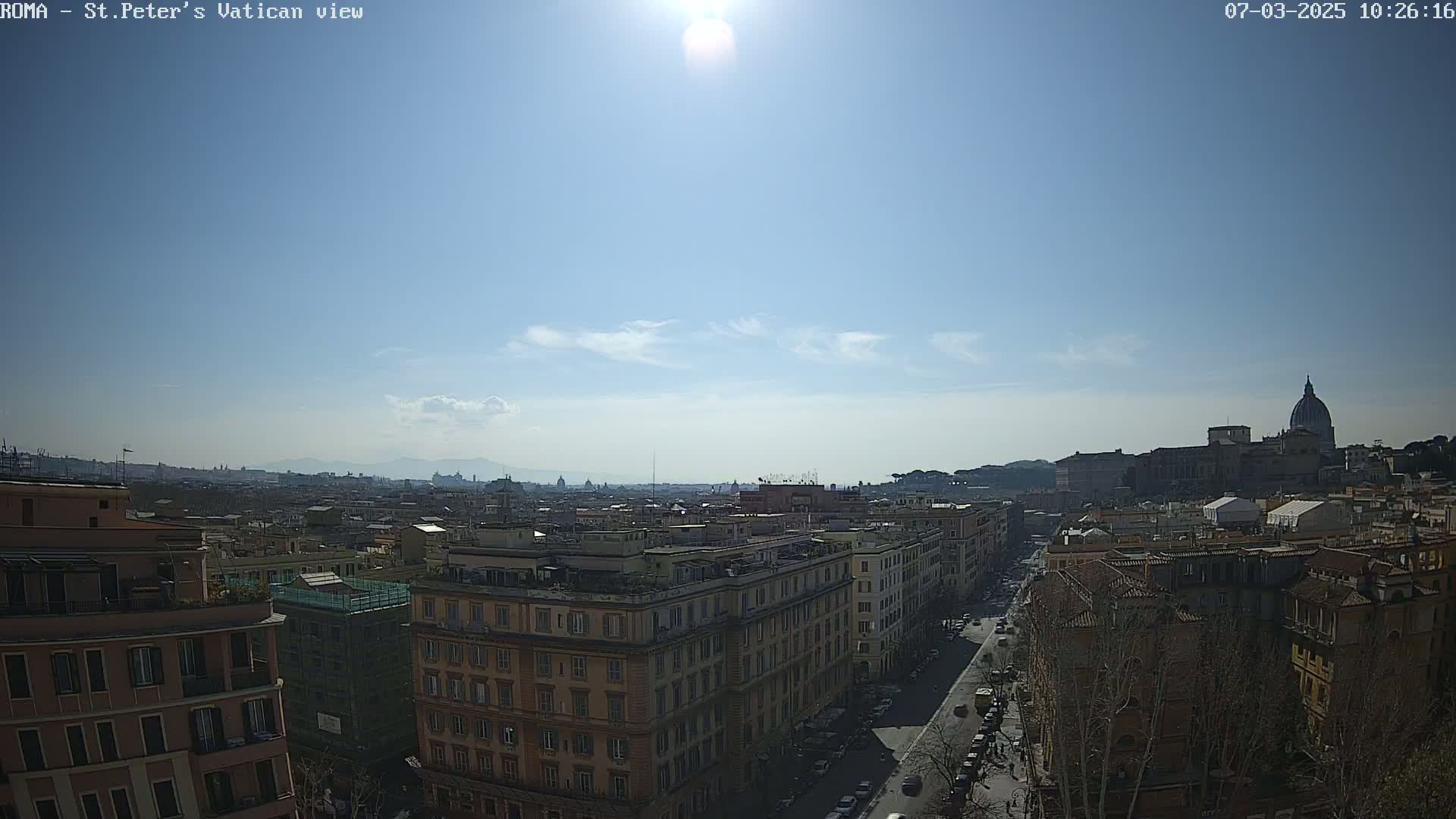 A sunny cityscape view shows a street lined with buildings leading towards St. Peter's Basilica in the distance.