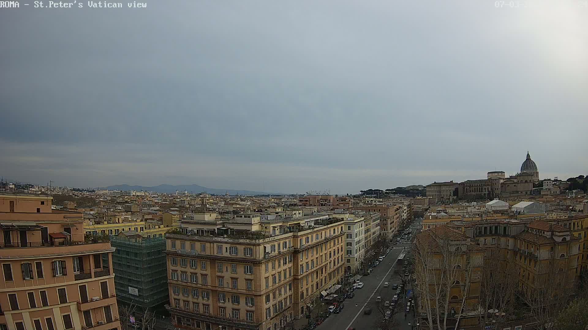A high-angle view of a cityscape under an overcast sky, showing a distant basilica with a dome and many buildings stretching to the horizon.