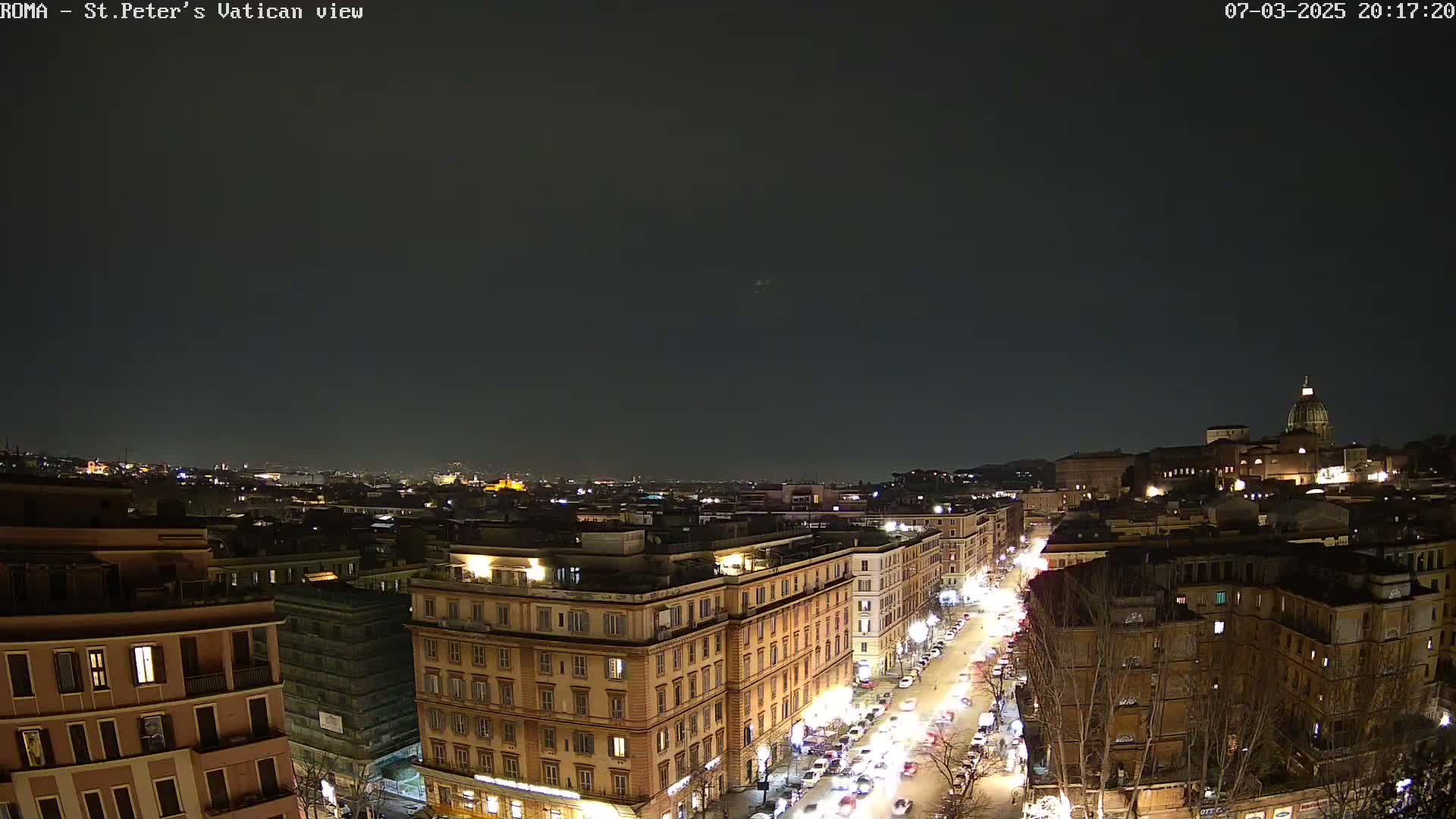 A nighttime aerial view of a city, showing illuminated buildings and streets with cars, under a dark, clear sky.