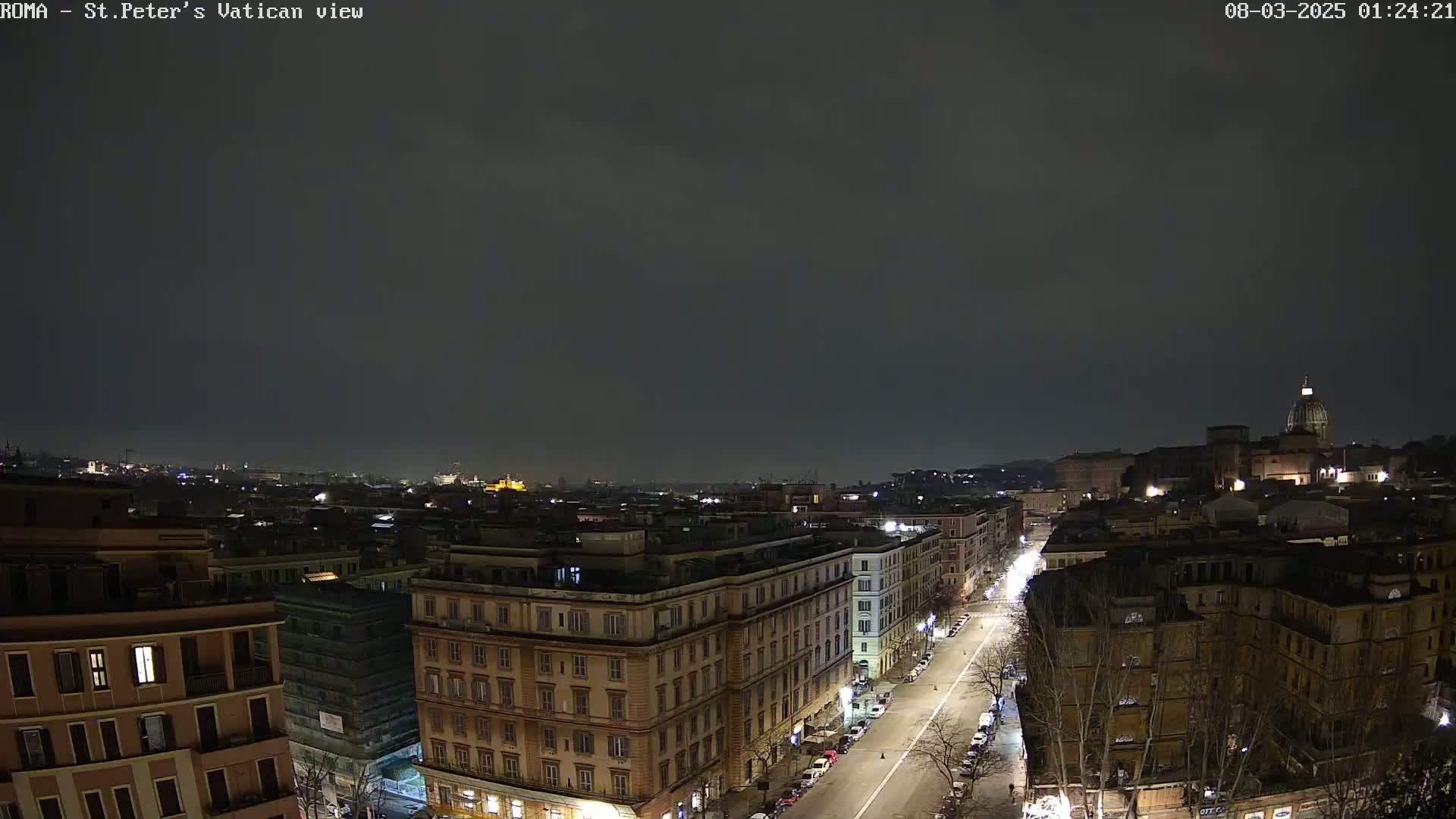 A nighttime, overcast view of a city, showing a long street lined with buildings leading toward a large dome-topped structure in the distance.