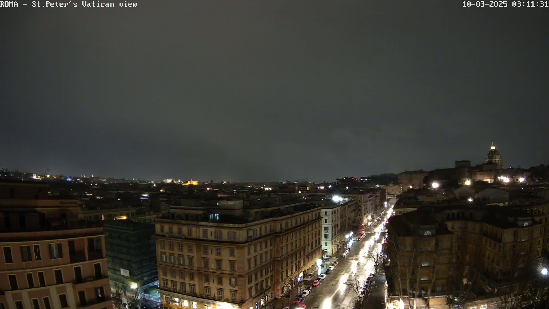 A nighttime, overcast view of a city street scene showing several buildings and illuminated vehicles under a dark sky.