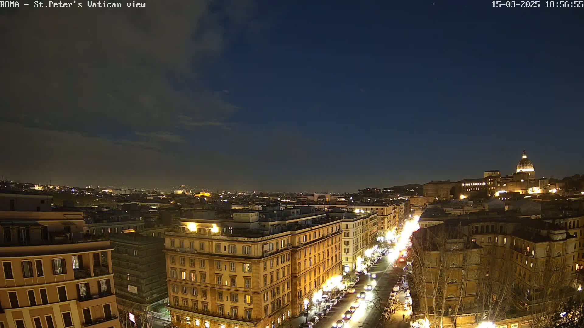 A nighttime view of a city, possibly Rome, shows illuminated buildings and streets, with a dome-shaped structure visible in the distance under a mostly clear, dark blue sky.
