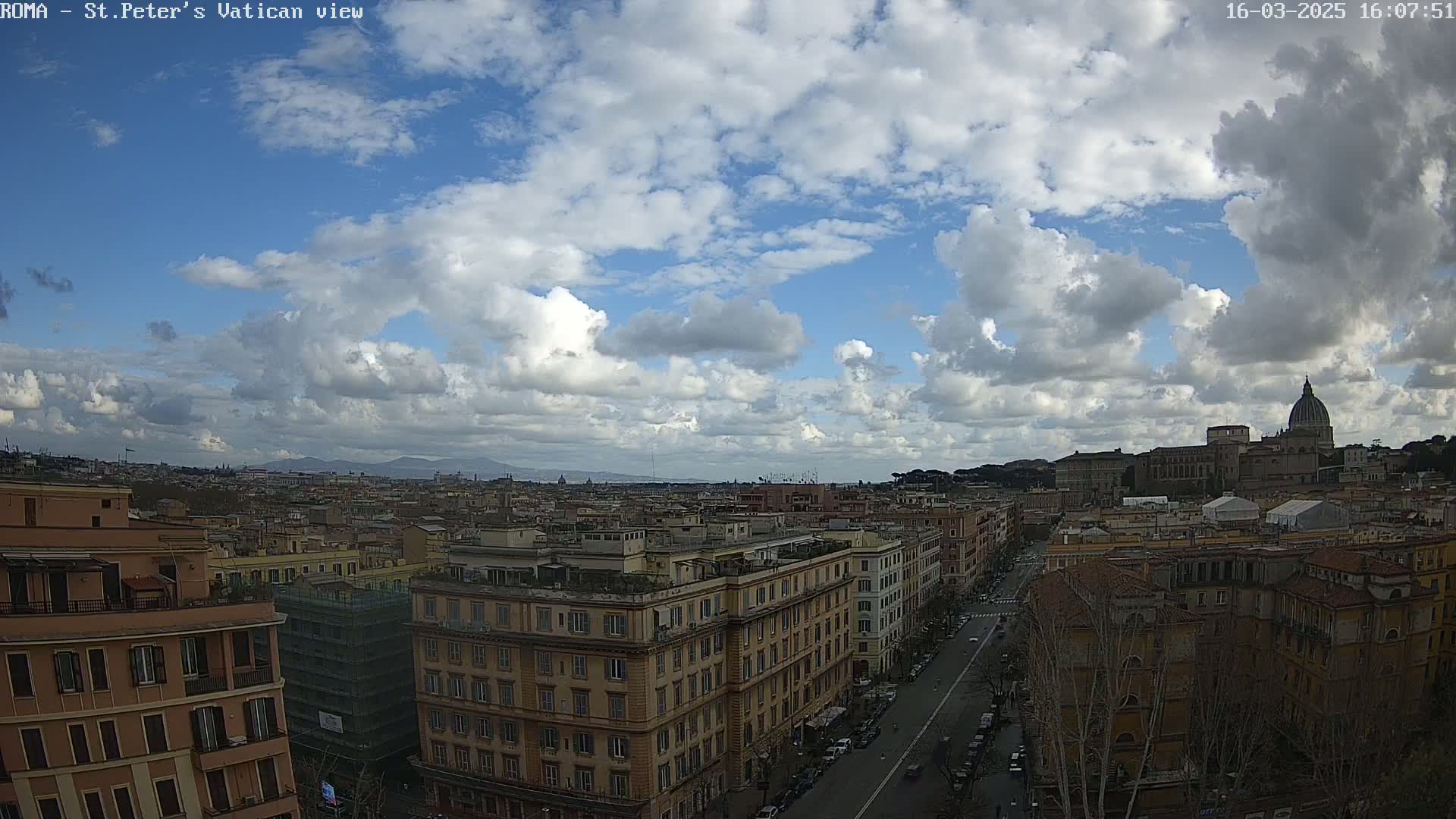 A partly cloudy sky overlooks a cityscape of multi-story buildings and a street with parked cars, featuring a large domed structure in the distance.