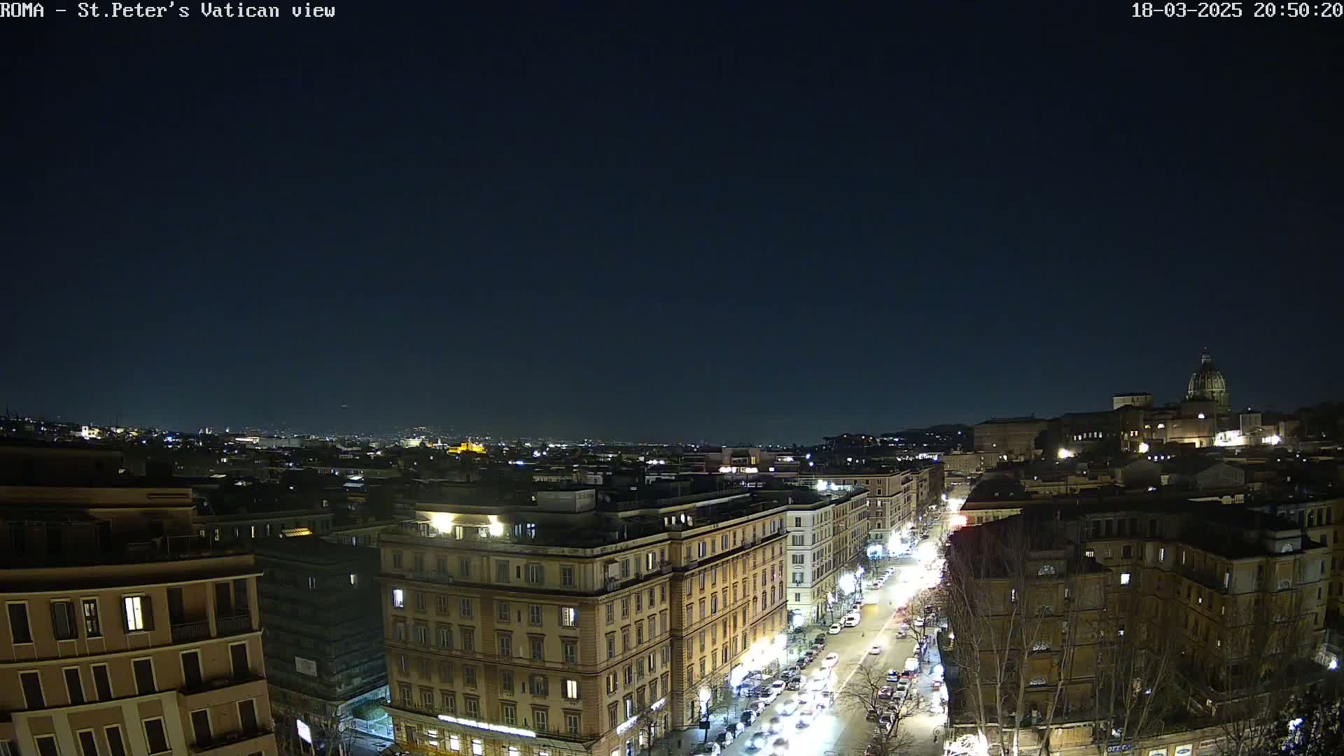 A nighttime, high-angle view of a city, showing buildings and a street with traffic under a clear, dark sky.