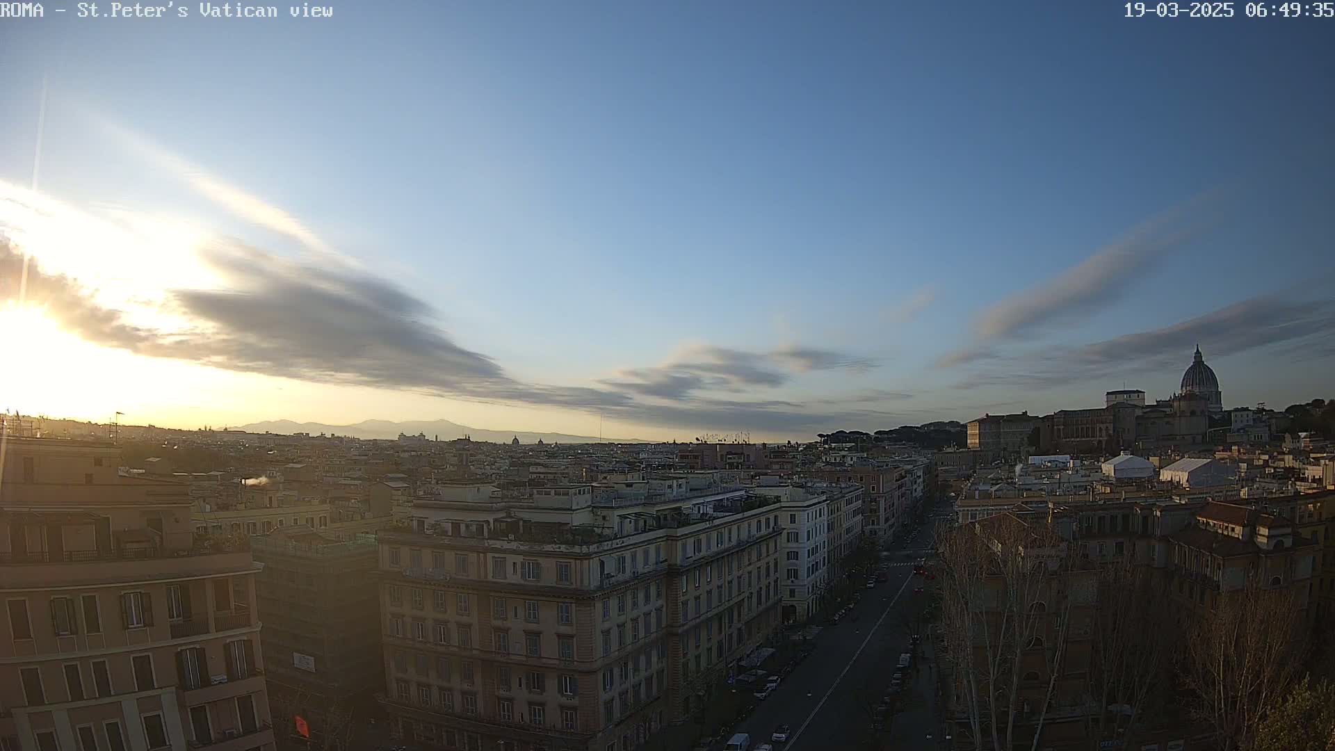 A cityscape at sunrise, with a partly cloudy sky, shows a view of buildings and St. Peter's Basilica in the distance.