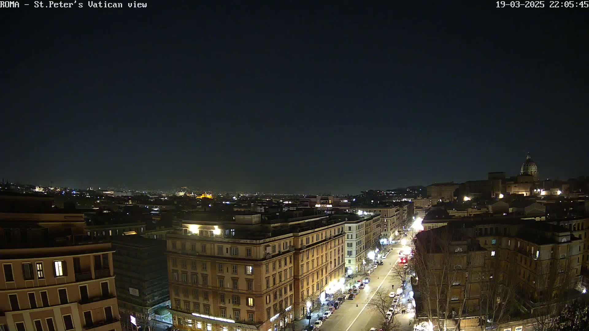 A nighttime, aerial view of a city shows a street lined with buildings, with a dome-shaped structure visible in the distance under a clear, dark sky.