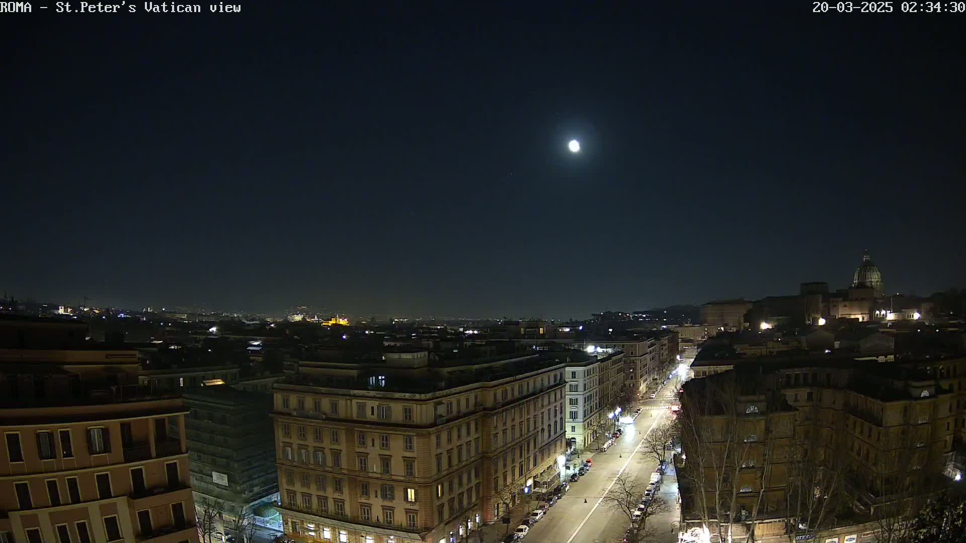 A nighttime, moonlit view of a city street lined with buildings, with a larger structure and dome visible in the distance.