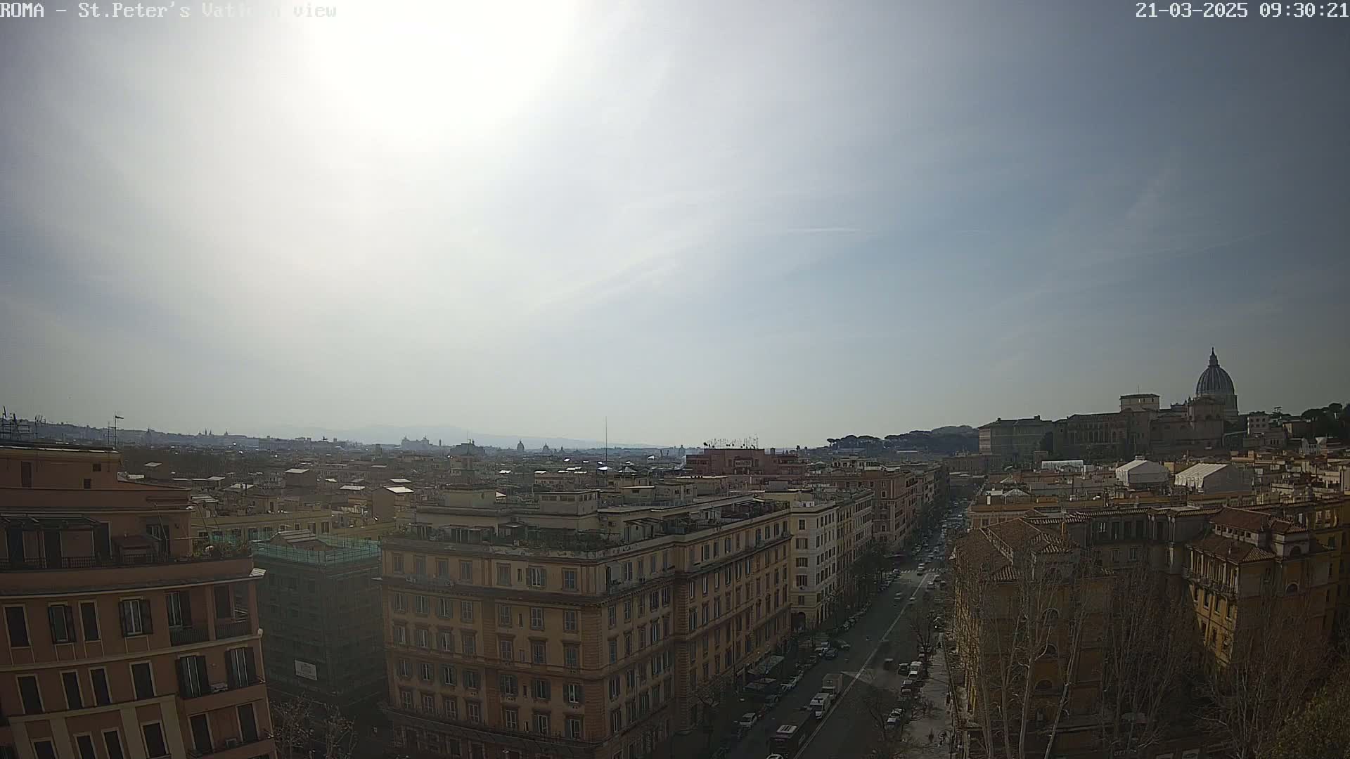 A sunny cityscape view shows a long street lined with buildings leading to a large domed structure in the distance.