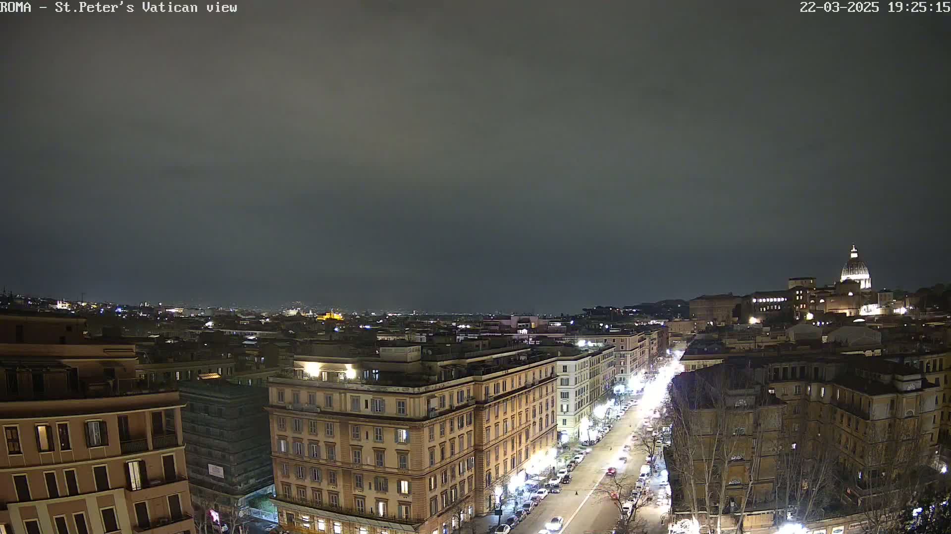 A nighttime, high-angle view of a city street scene shows illuminated buildings, cars, and trees under a dark, cloudy sky.