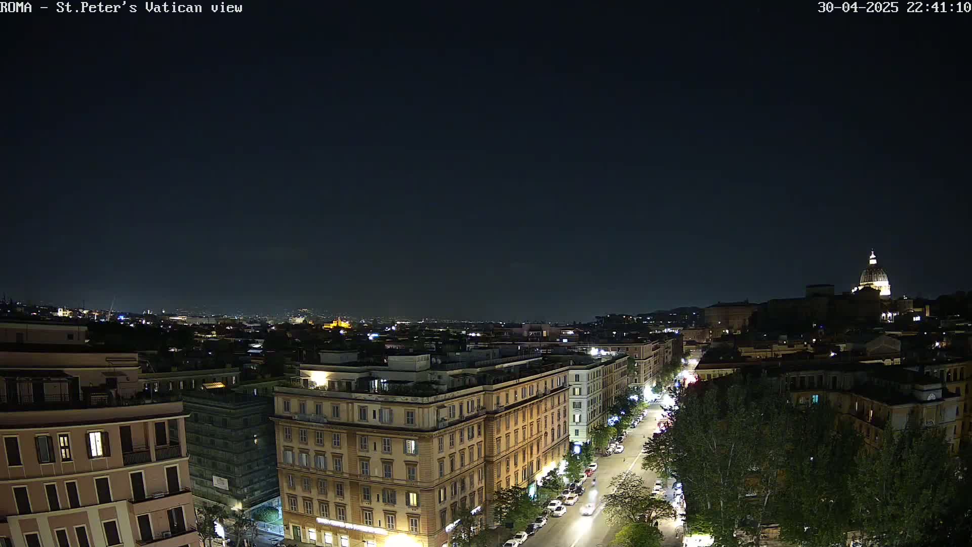A nighttime aerial view of a city, showing a street lined with buildings and trees, with a large dome in the distance under a clear, dark sky.