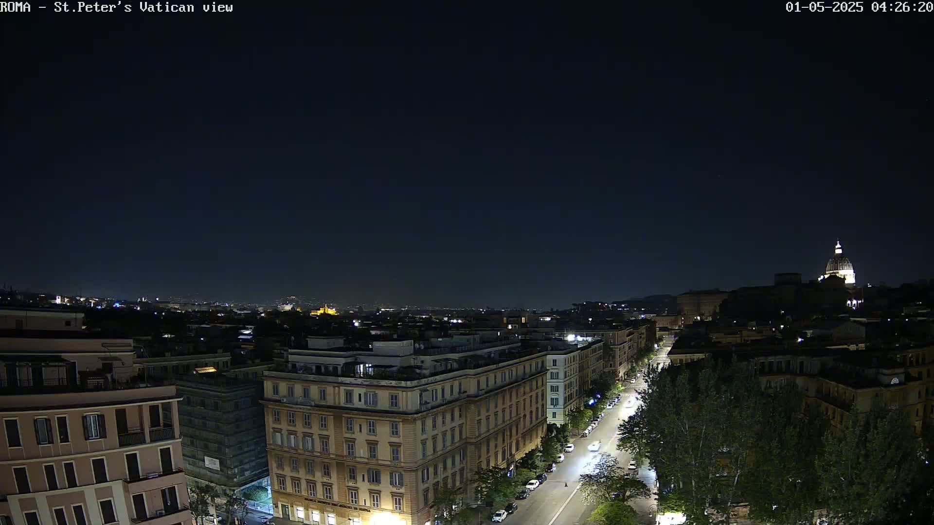 A nighttime cityscape shows a long street lined with buildings and trees leading toward a dome-shaped structure in the distance under a clear, dark sky.