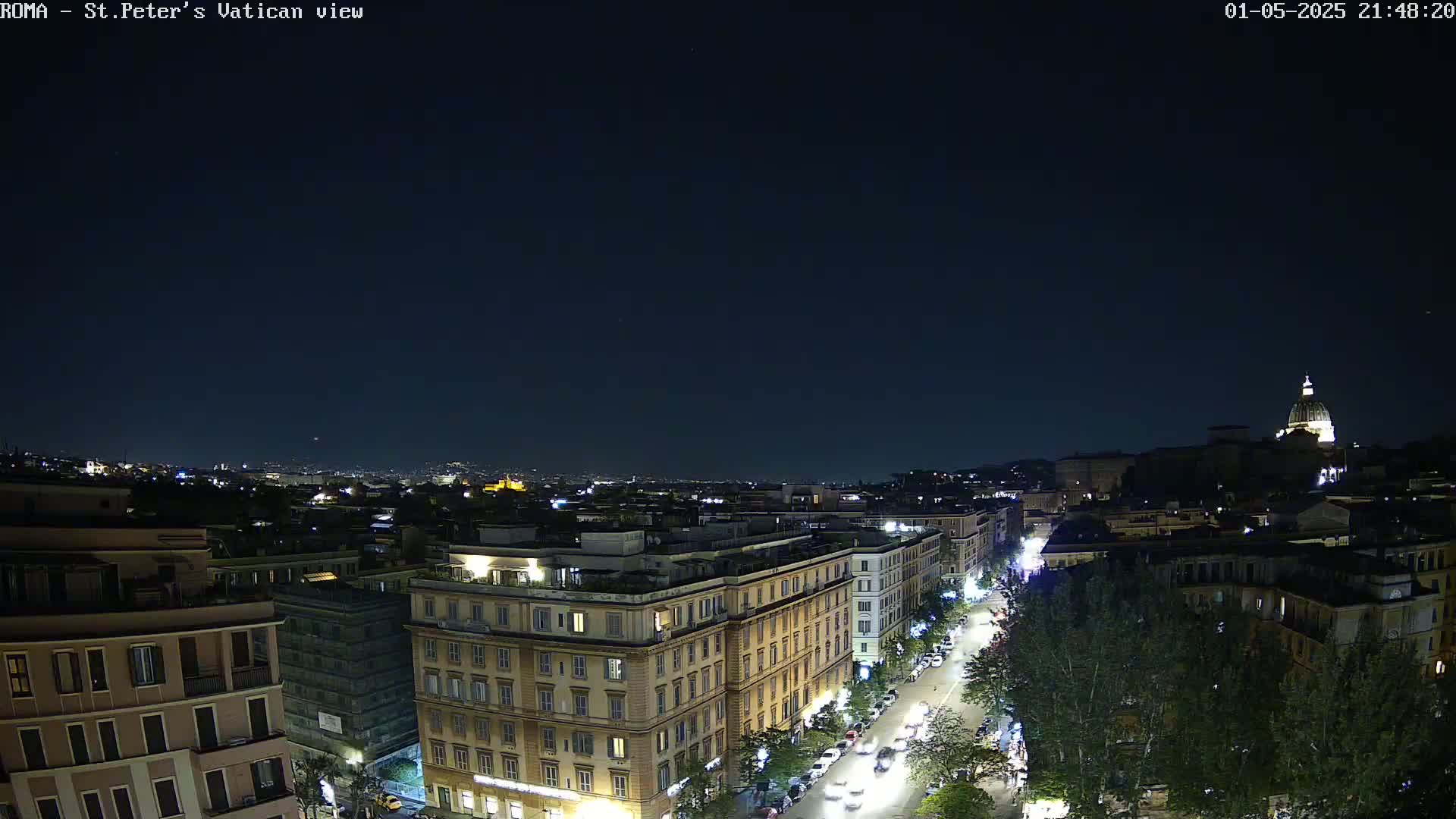 A nighttime, high-angle view of a city, showing buildings, streets with cars, and trees, with a large dome visible in the distance under a clear, dark sky.