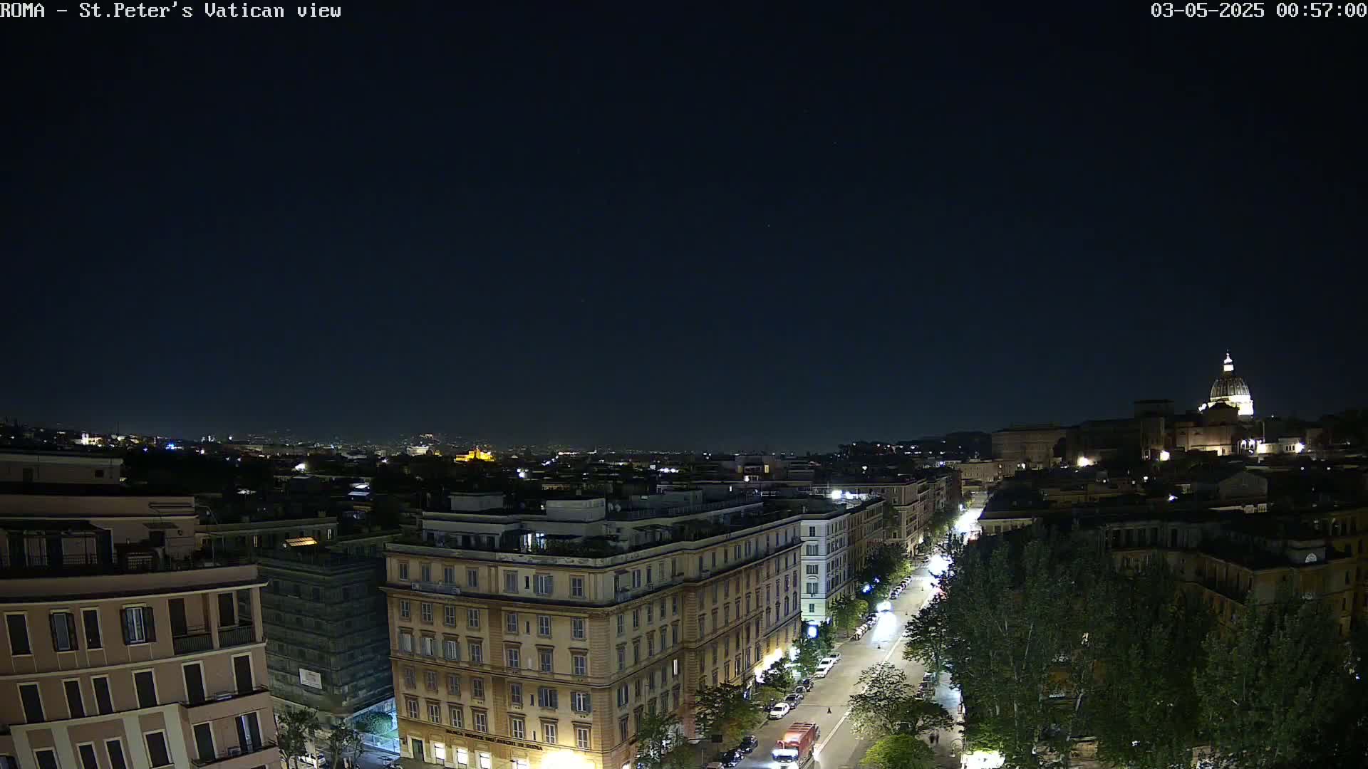 A nighttime, aerial view of a city street lined with trees and buildings, with a distant dome-shaped structure illuminated under a clear, dark sky.