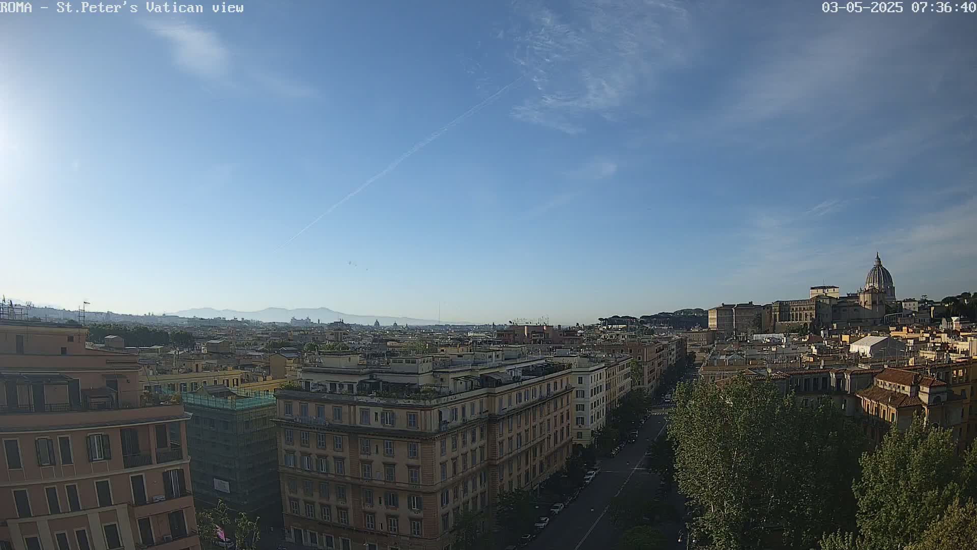 A mostly clear, sunny day reveals a panoramic view of Rome's cityscape, including multi-story buildings, a tree-lined street, and the dome of St. Peter's Basilica in the distance.
