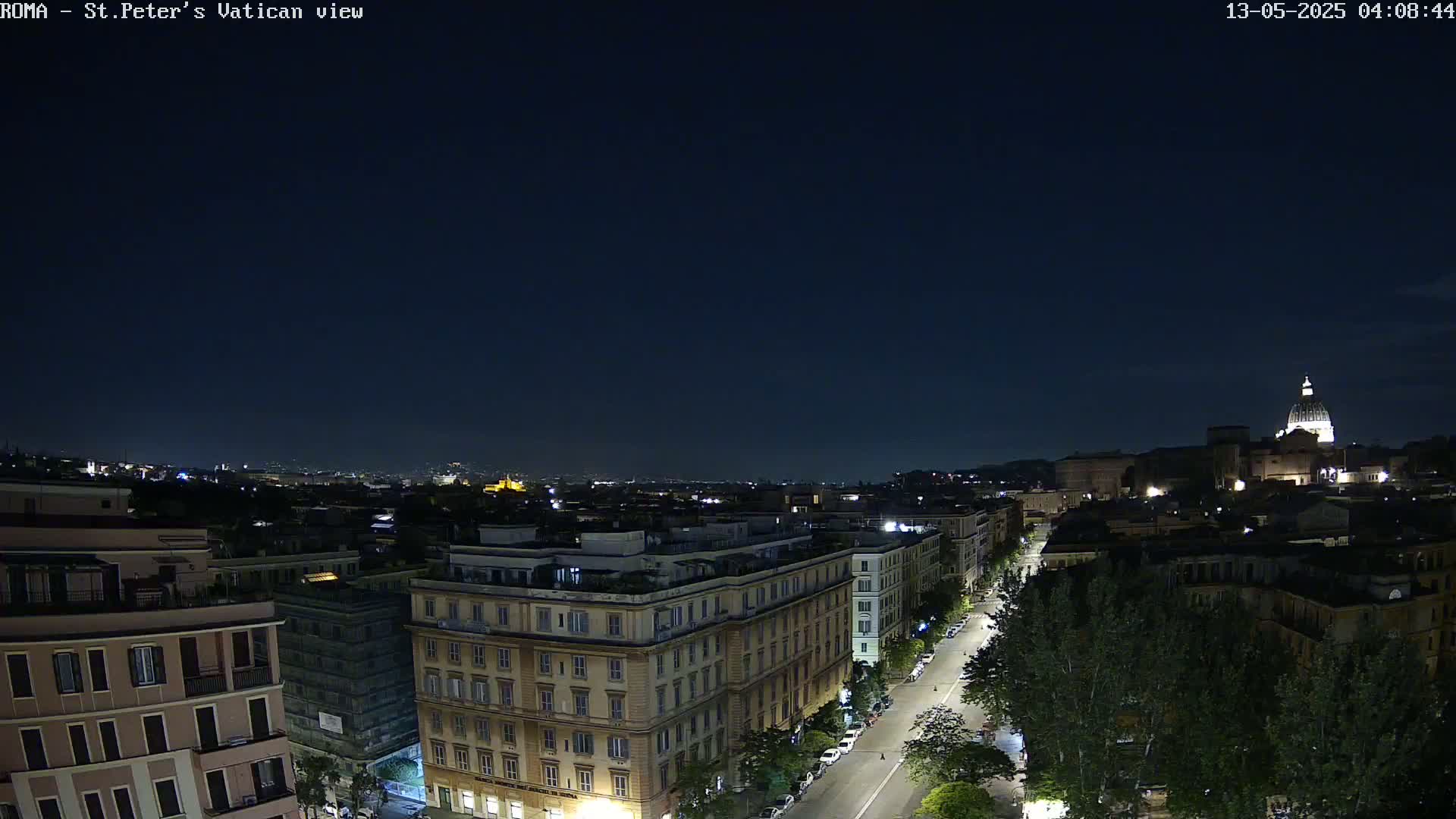 A nighttime cityscape view shows a city street lined with buildings, trees, and parked cars, extending towards a distant structure with a dome under a clear, dark sky.