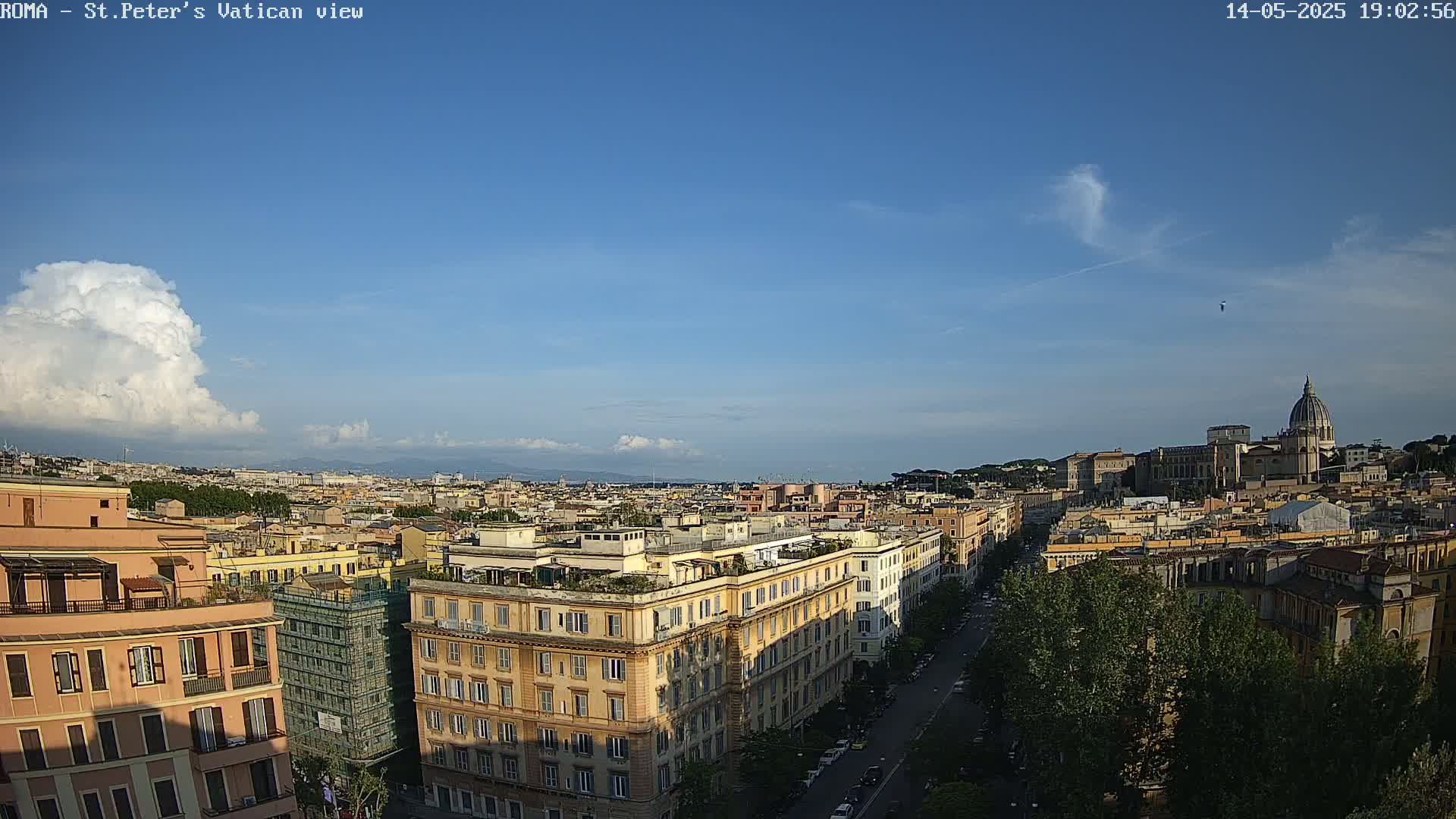 A partly sunny cityscape view of Rome, Italy, shows a distant St. Peter's Basilica and numerous buildings under a mostly clear blue sky with a single large cumulus cloud.