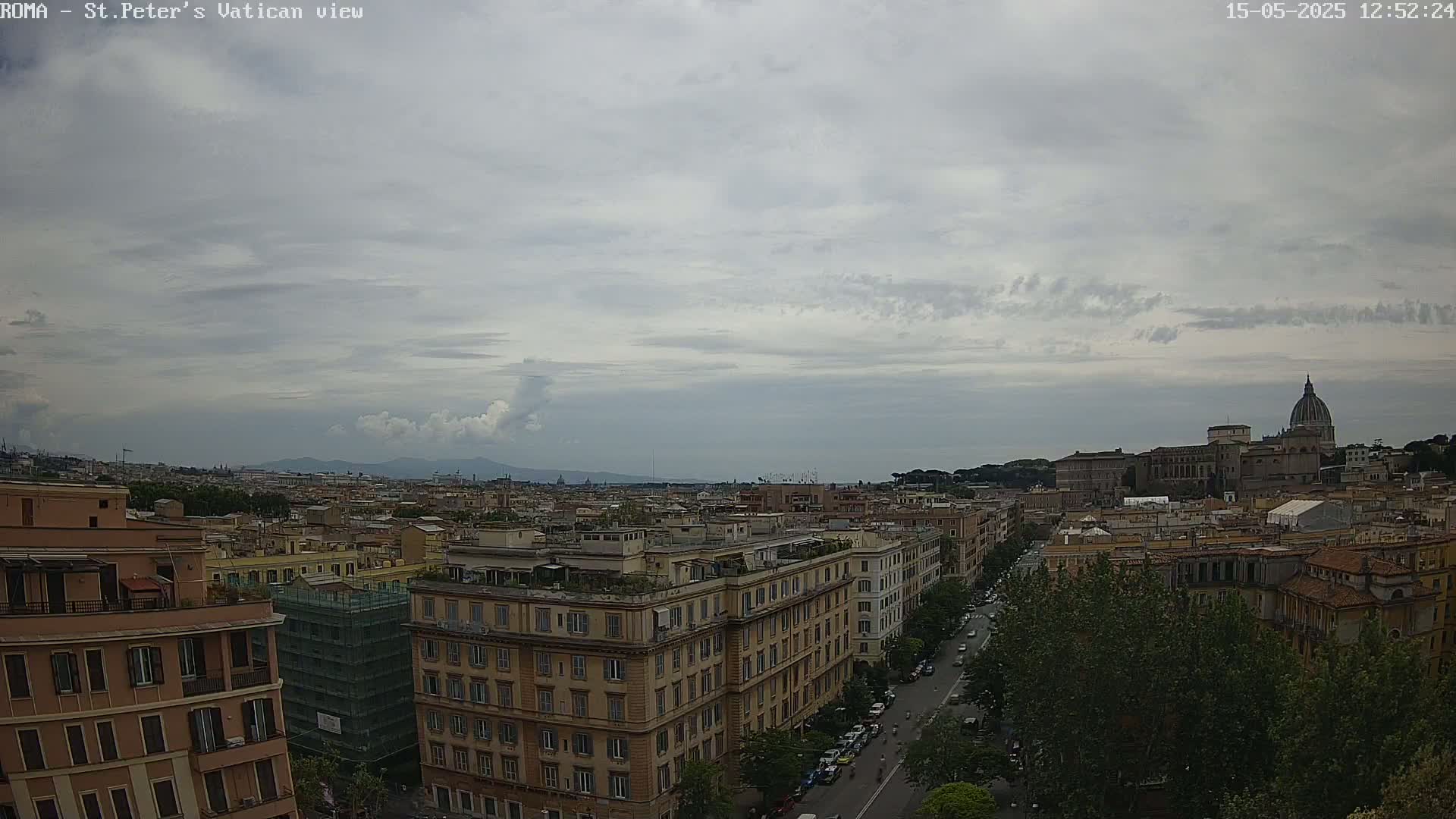 A high-angle view of Rome on an overcast day shows a cityscape with buildings, trees, and the dome of St. Peter's Basilica in the distance.