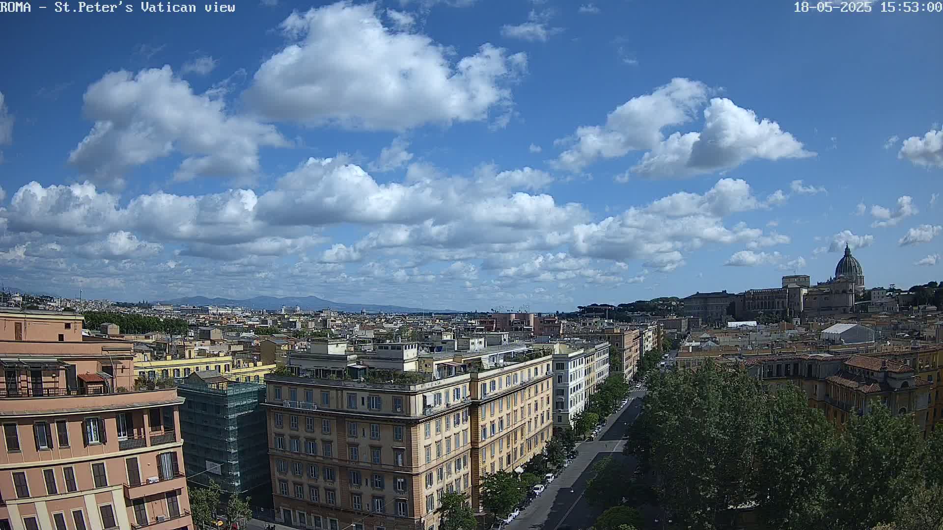A sunny day with partly cloudy skies overlooks a cityscape featuring several multi-story buildings, a street lined with trees, and a prominent domed structure in the distance.