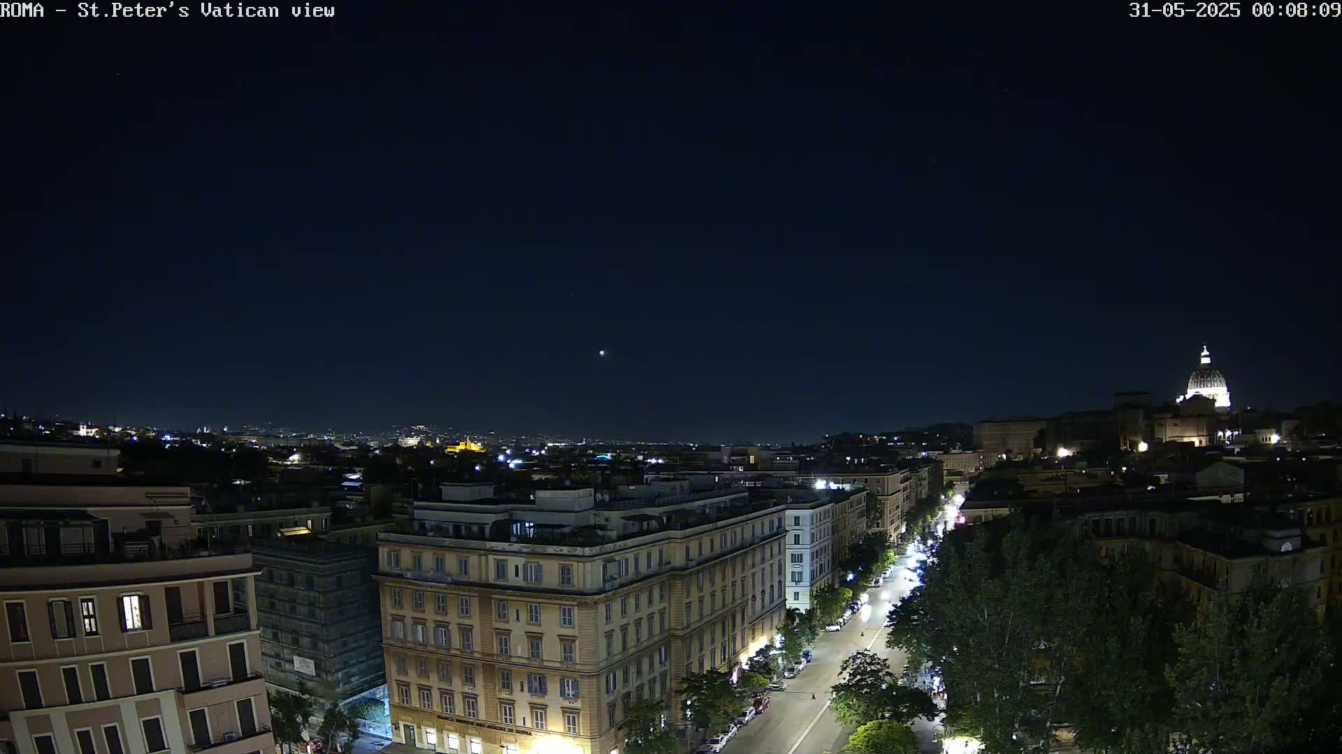 A nighttime aerial view of a city street lined with trees and buildings, with the dome of a large building visible in the distance under a clear, dark sky.