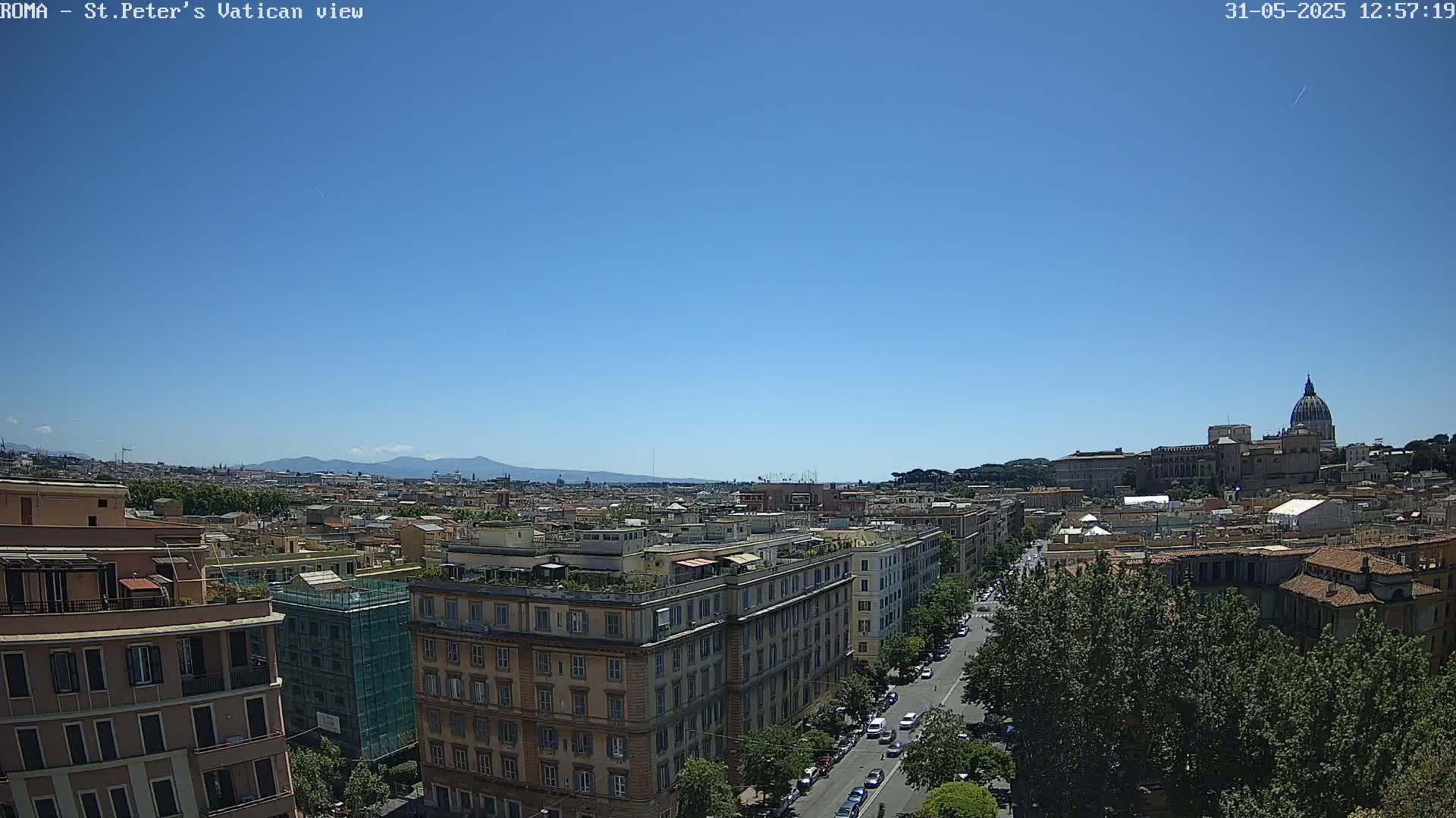 A high-angle view of Rome on a clear, sunny day shows a cityscape stretching to distant mountains, with St. Peter's Basilica visible in the background.