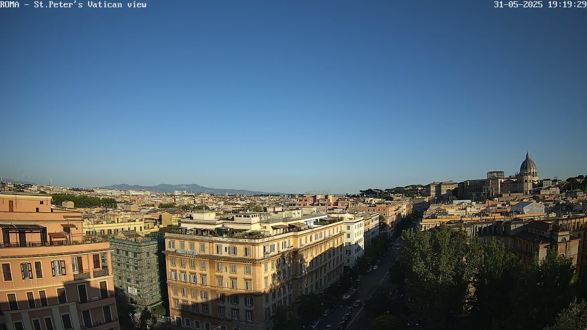 A high-angle, long shot of a cityscape under a clear blue sky shows buildings and streets stretching to the horizon, with a large domed structure visible in the distance.