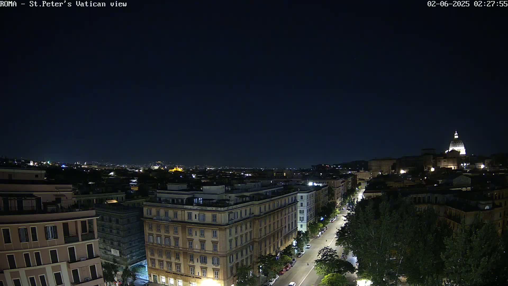 A nighttime view of a city street lined with buildings, with the dome of a large structure visible in the distance under a clear, dark sky.