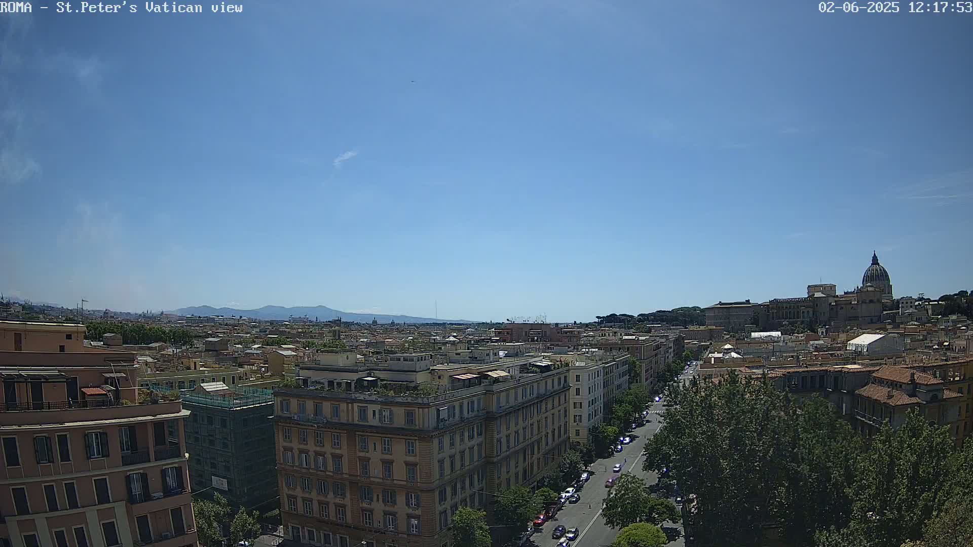 A high-angle, long shot on a clear, sunny day shows a cityscape with the dome of St. Peter's Basilica visible in the distance, and mountains on the horizon.