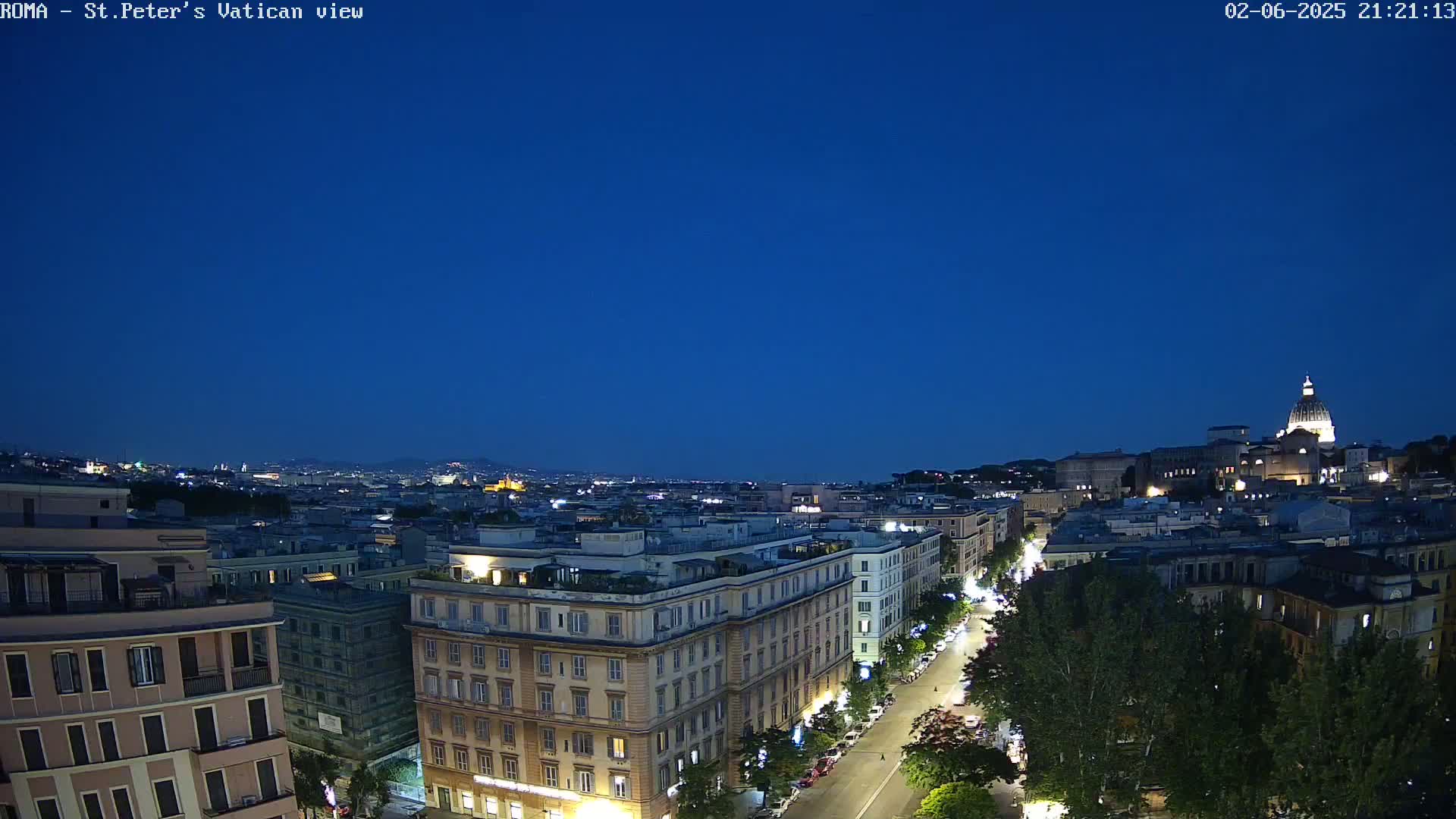A nighttime aerial view of a city, showing buildings illuminated against a dark blue sky, with a large dome-shaped structure visible in the distance.