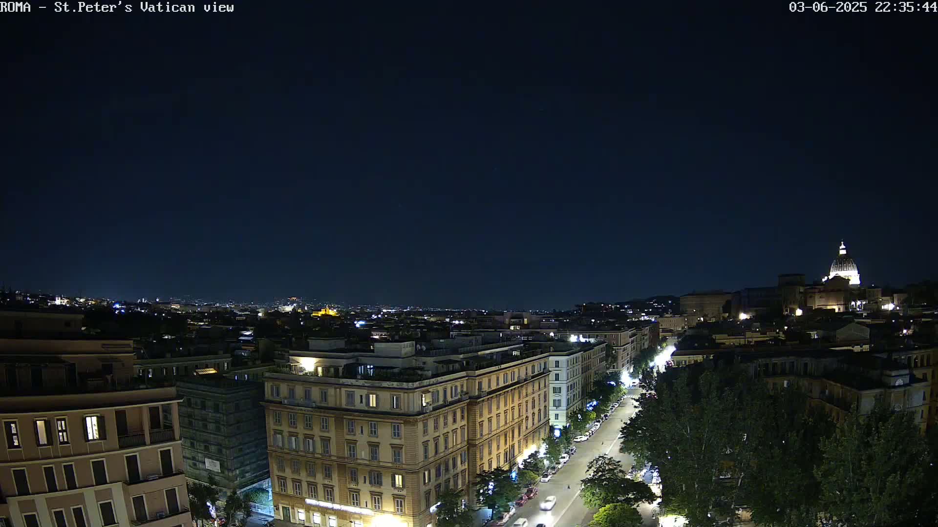 A nighttime, aerial view of a city, showing a street lined with trees and buildings, with a dome-shaped structure visible in the distance under a clear, dark sky.