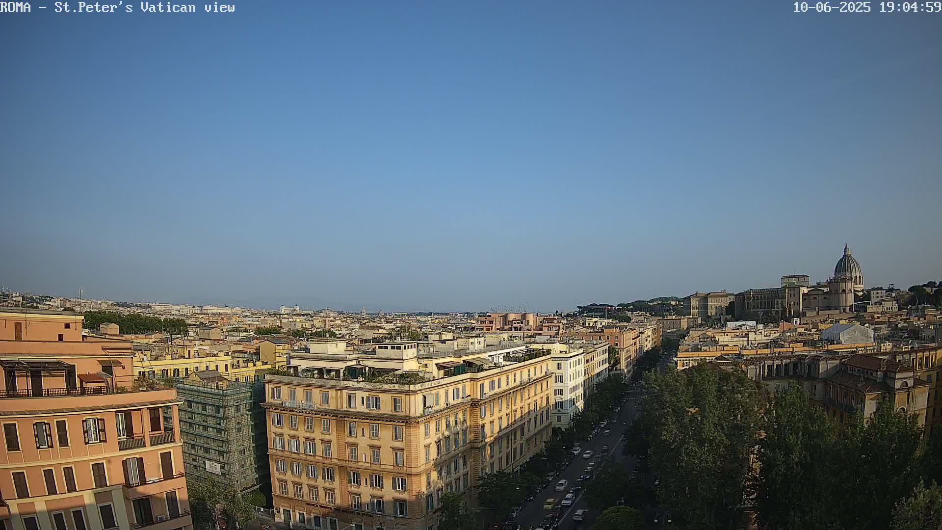 A high-angle, long shot of a city under a clear blue sky shows a cityscape with buildings and a large dome in the distance.