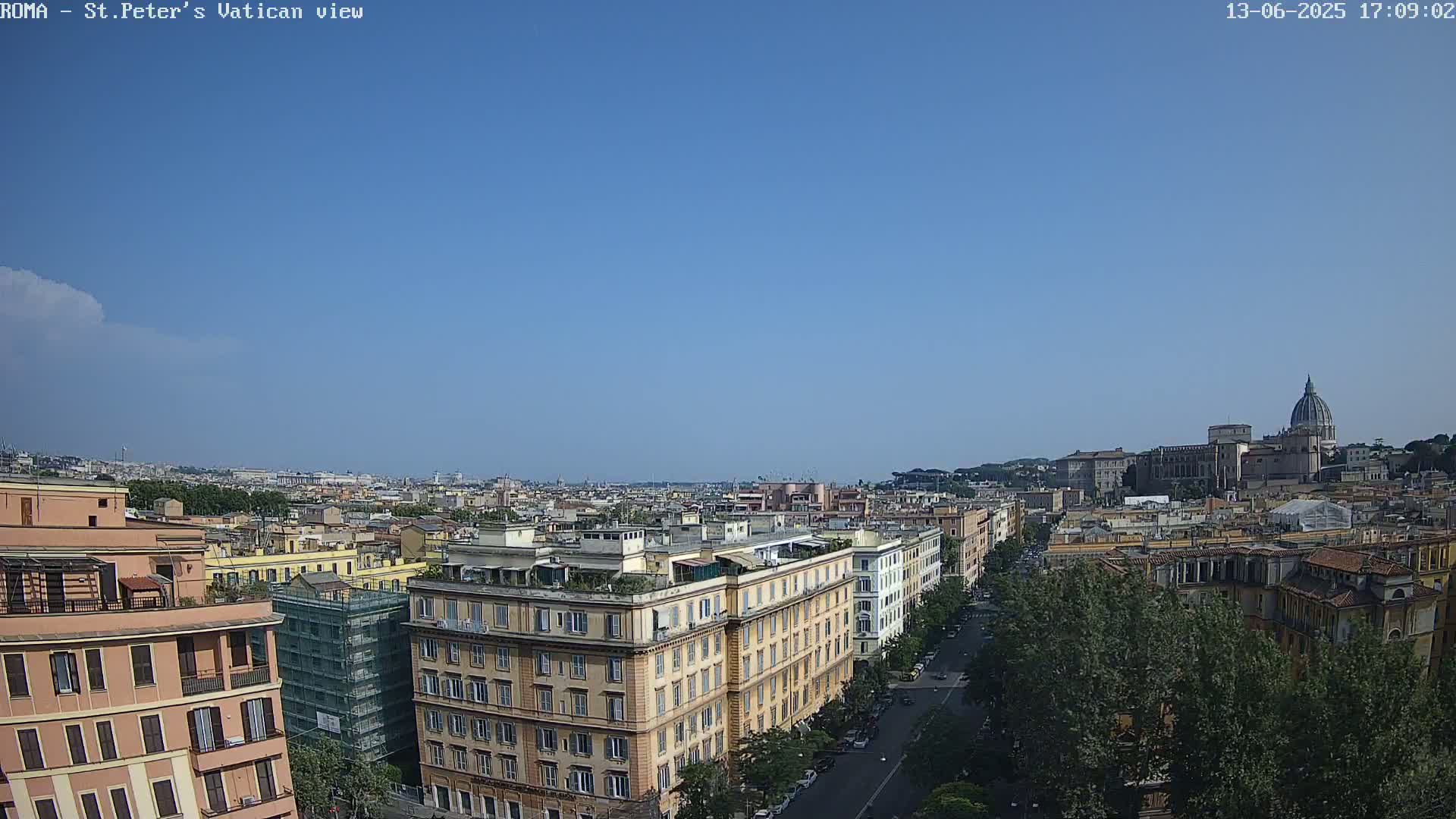 A wide, high-angle view of a city on a clear, sunny day shows numerous buildings stretching to the horizon, with St. Peter's Basilica visible in the distance.