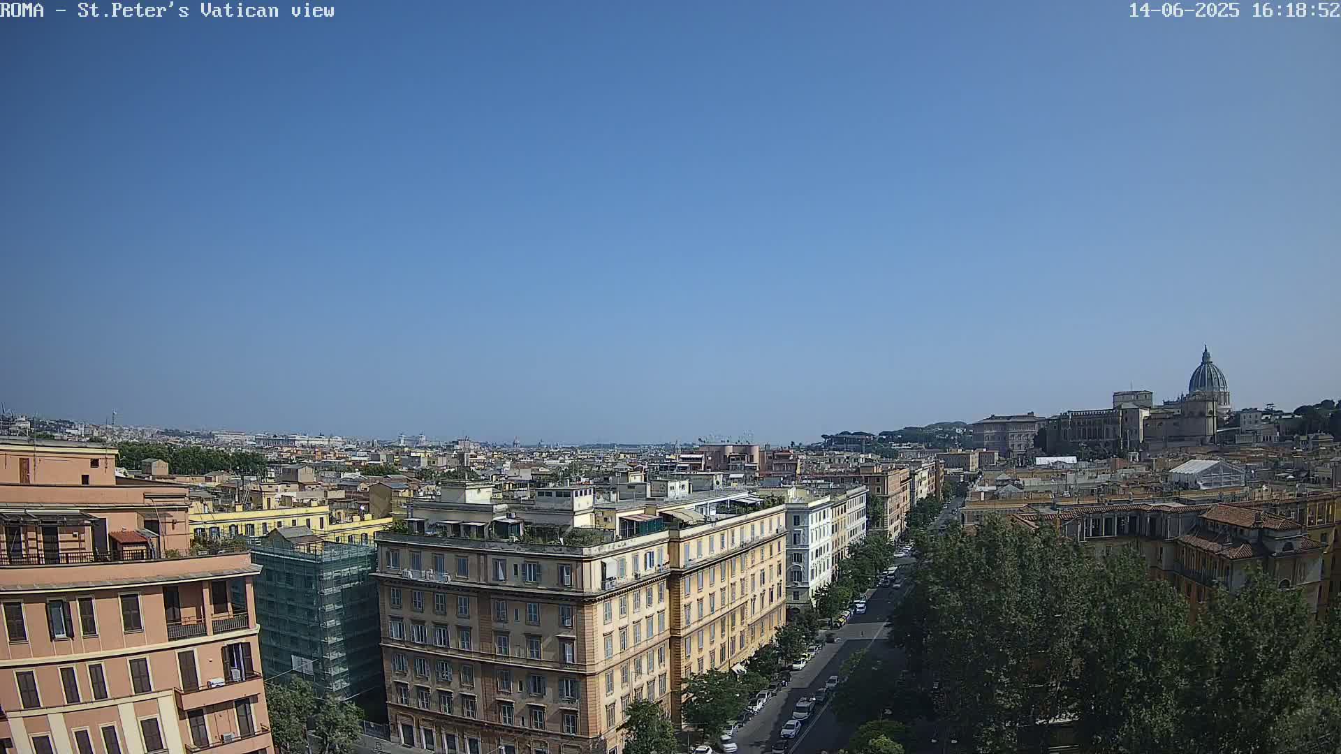 A high-angle, long shot on a clear, sunny day shows a cityscape with numerous buildings, a tree-lined street, and the dome of a large church in the distance.