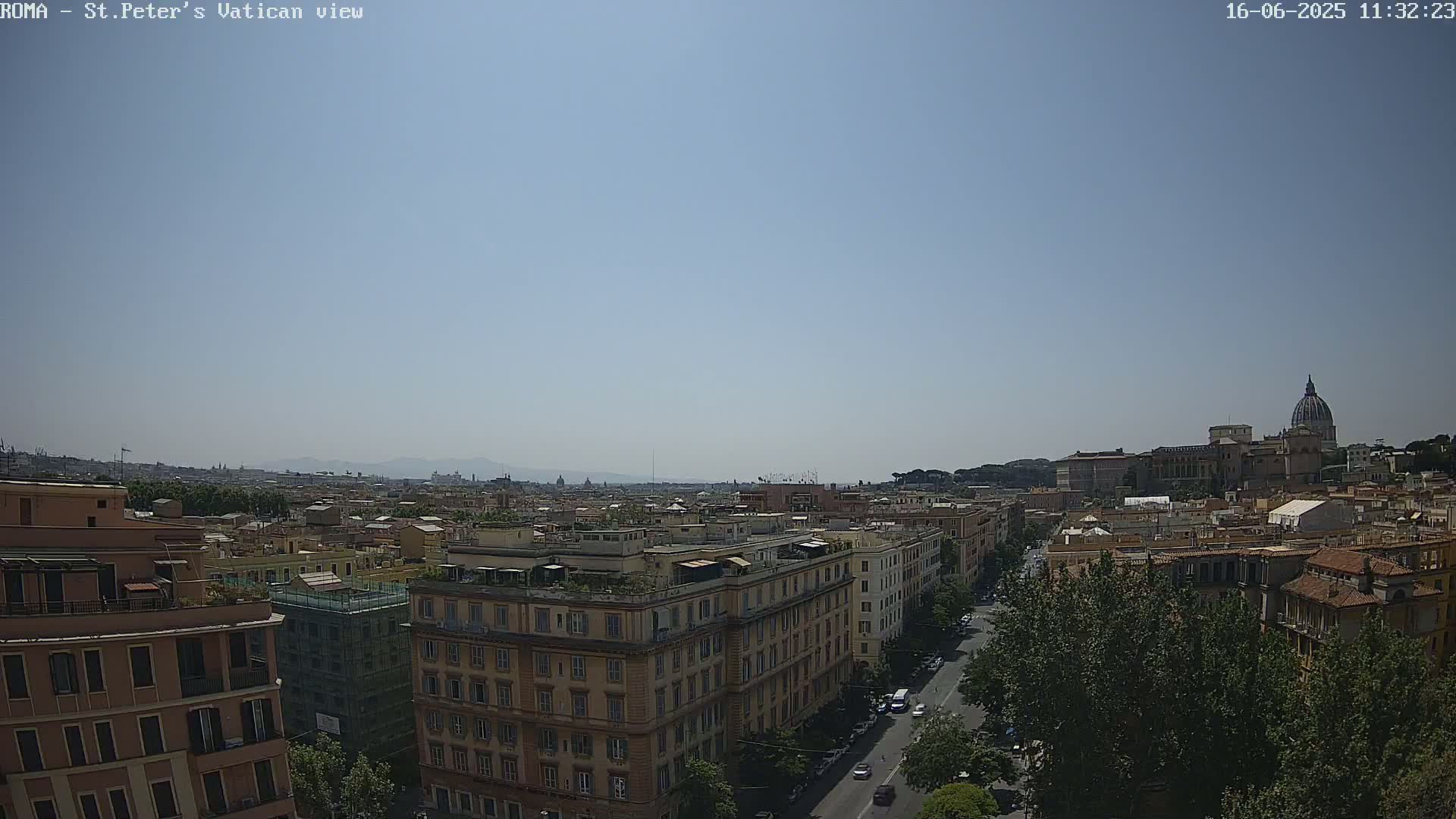 A high-angle, long shot on a clear, sunny day shows a cityscape with multi-story buildings, a street with cars, and the dome of St. Peter's Basilica in the distance.
