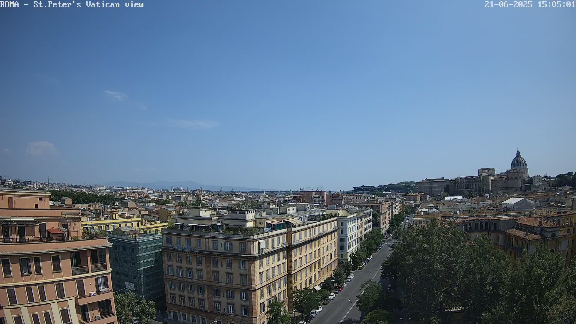 A wide shot of a city on a clear, sunny day shows a cityscape with multi-story buildings, a street, and trees, with a large domed building visible in the distance.