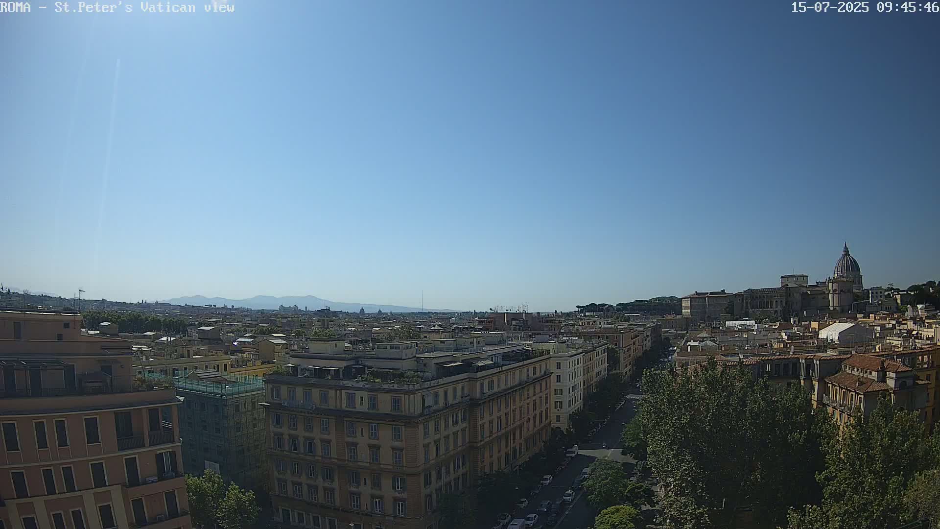 A clear, sunny day view of a cityscape, featuring multi-story buildings, a tree-lined street, and a large domed structure in the distance.
