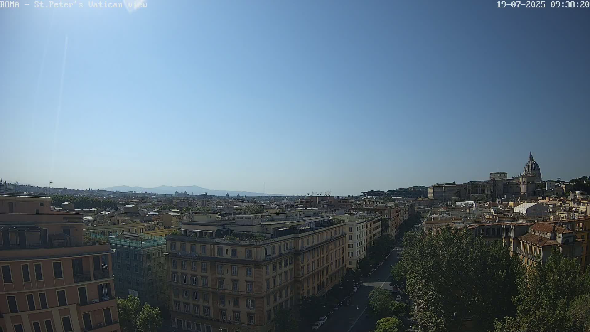 A high-angle, long shot on a clear, sunny day shows a cityscape with a large basilica in the distance, surrounded by buildings and trees.