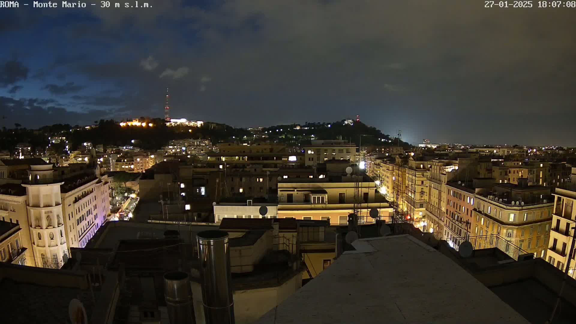 A nighttime view of a city, illuminated by streetlights and building lights, under a mostly cloudy sky.