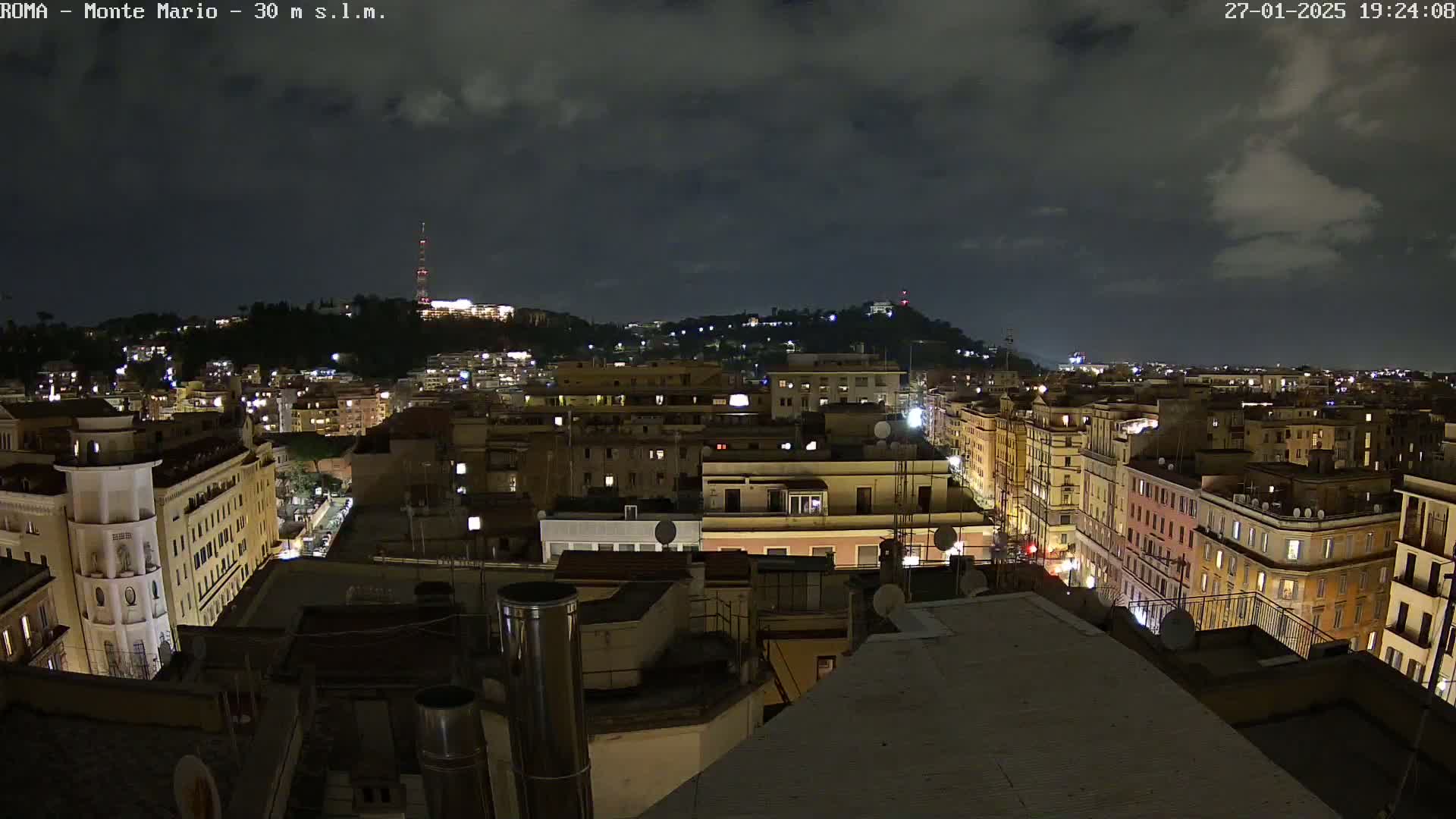 A nighttime view of a city with a hill in the background under a partly cloudy sky.