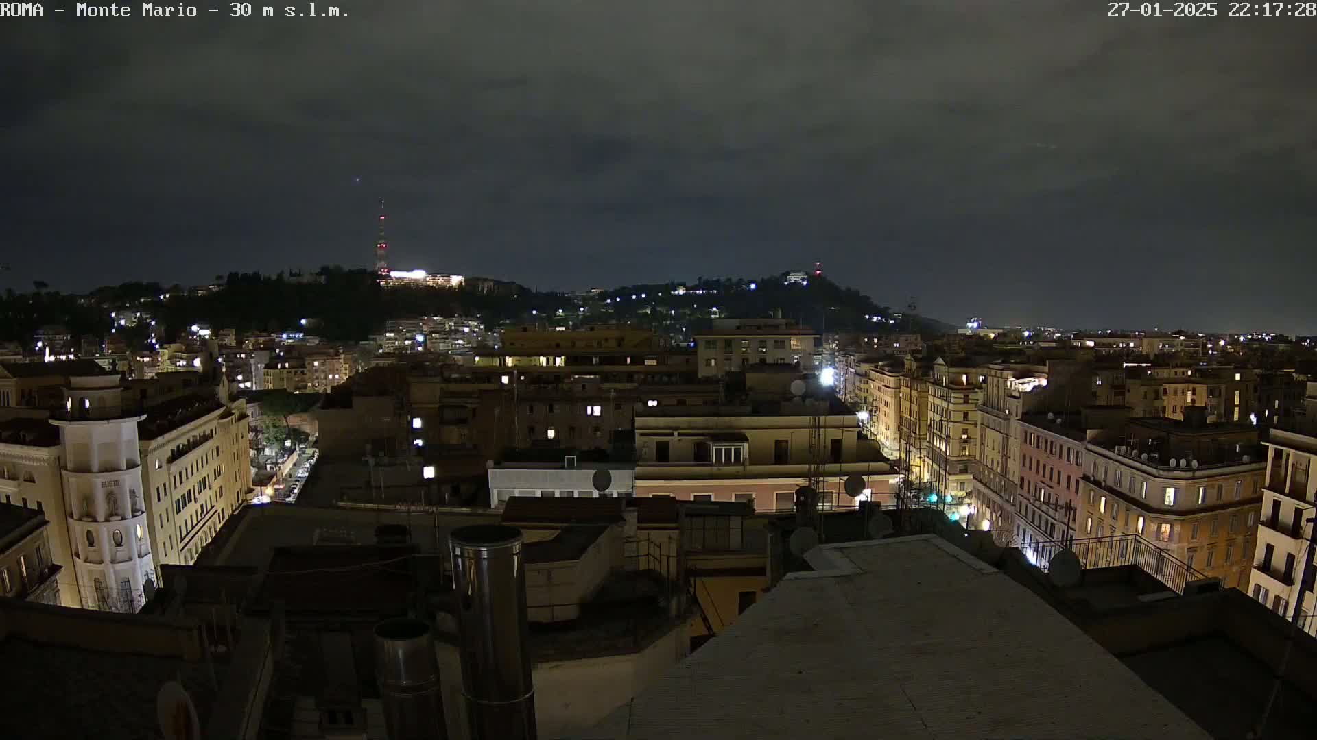 A nighttime view of a city, under a dark, cloudy sky, shows a cityscape of buildings illuminated by lights, with a hill in the background.