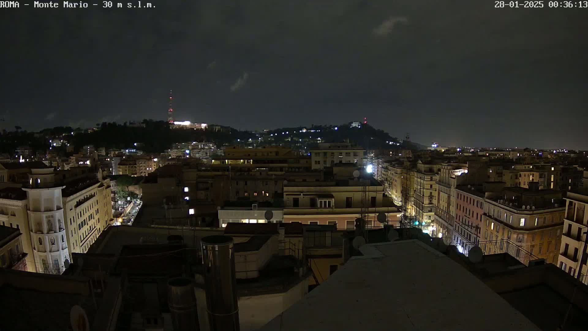 A nighttime view of a city with numerous buildings illuminated, under a dark, cloudy sky.