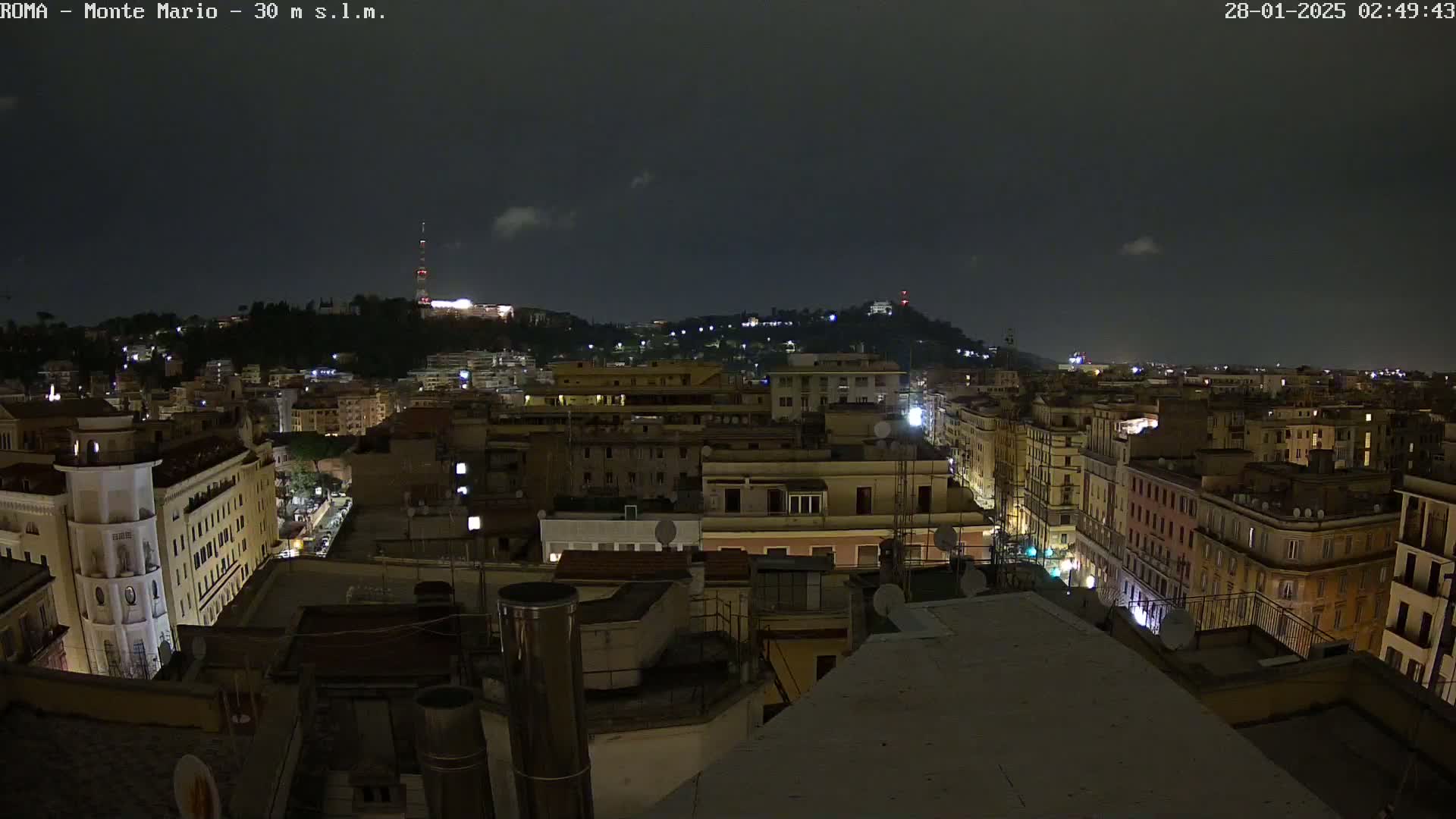 A nighttime, slightly cloudy view of a city, showing many buildings illuminated by streetlights and a hill in the background.