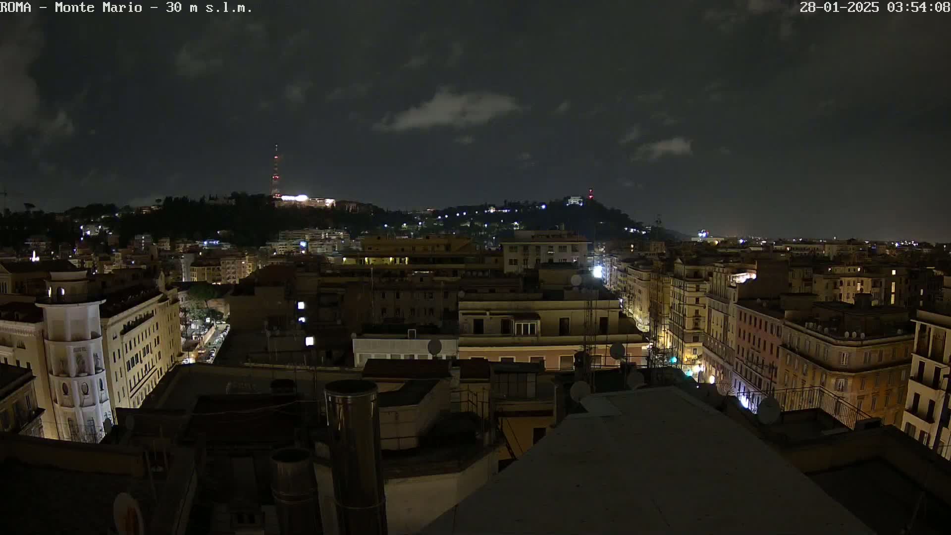 A nighttime cityscape with a partly cloudy sky shows many buildings illuminated, and a hill in the distance.