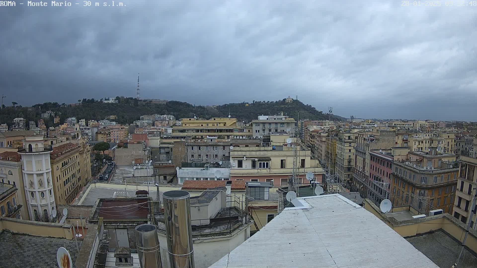 A rooftop view of a city under a cloudy sky shows numerous buildings extending to a hill in the distance.