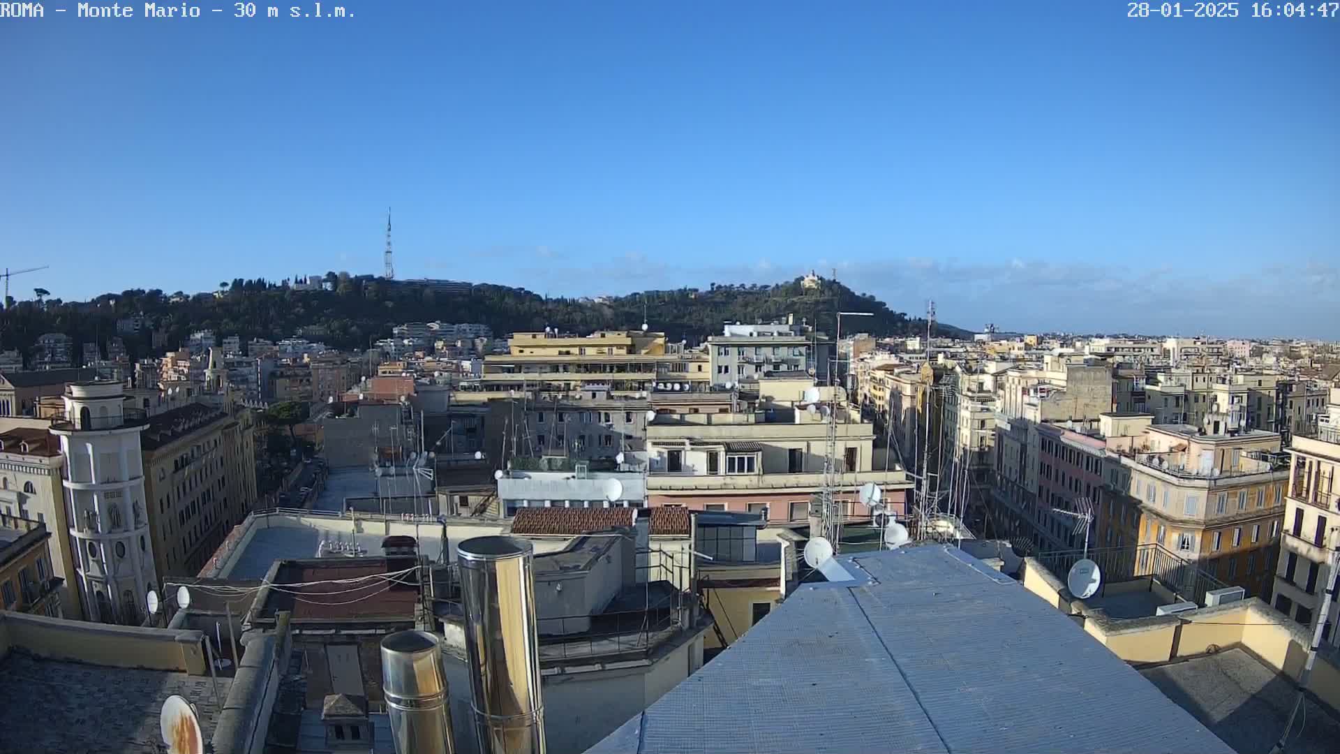 A high-angle, long shot on a clear, sunny day shows a cityscape of numerous buildings, with a hill in the background.