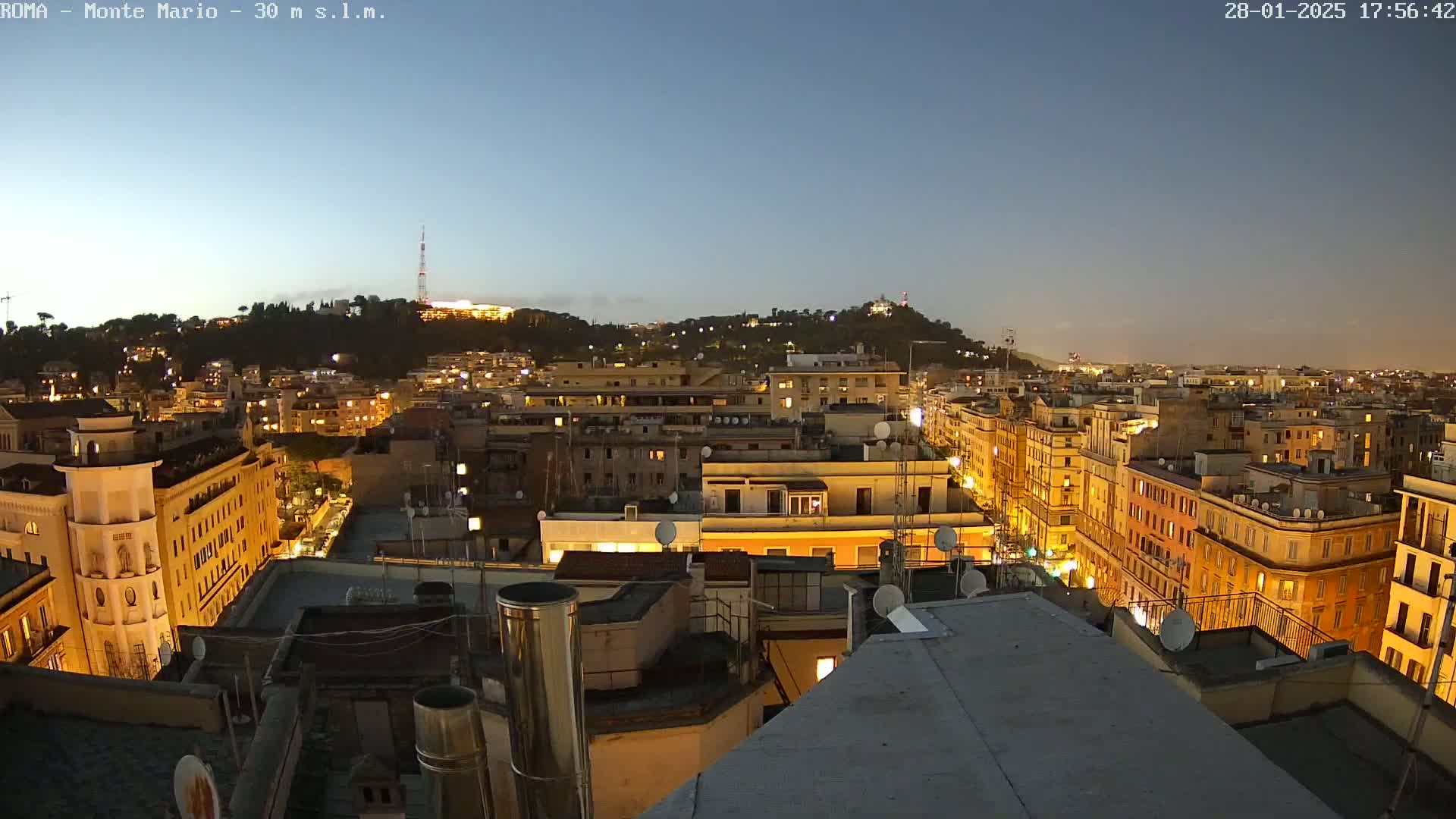 A nighttime view of a city with illuminated buildings and a hill in the background under a clear, twilight sky.