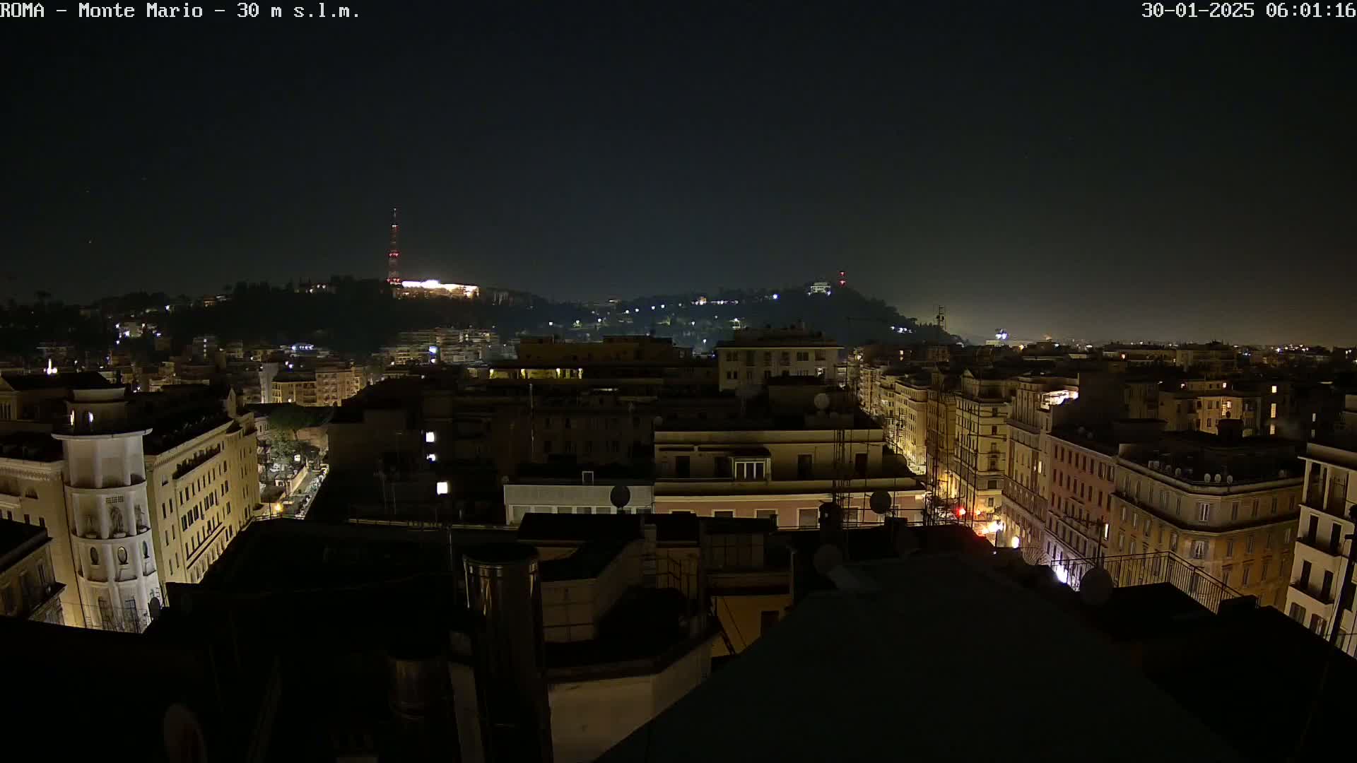 A nighttime cityscape view of a city with many buildings lit up, overlooking a hill in the distance under a clear, dark sky.