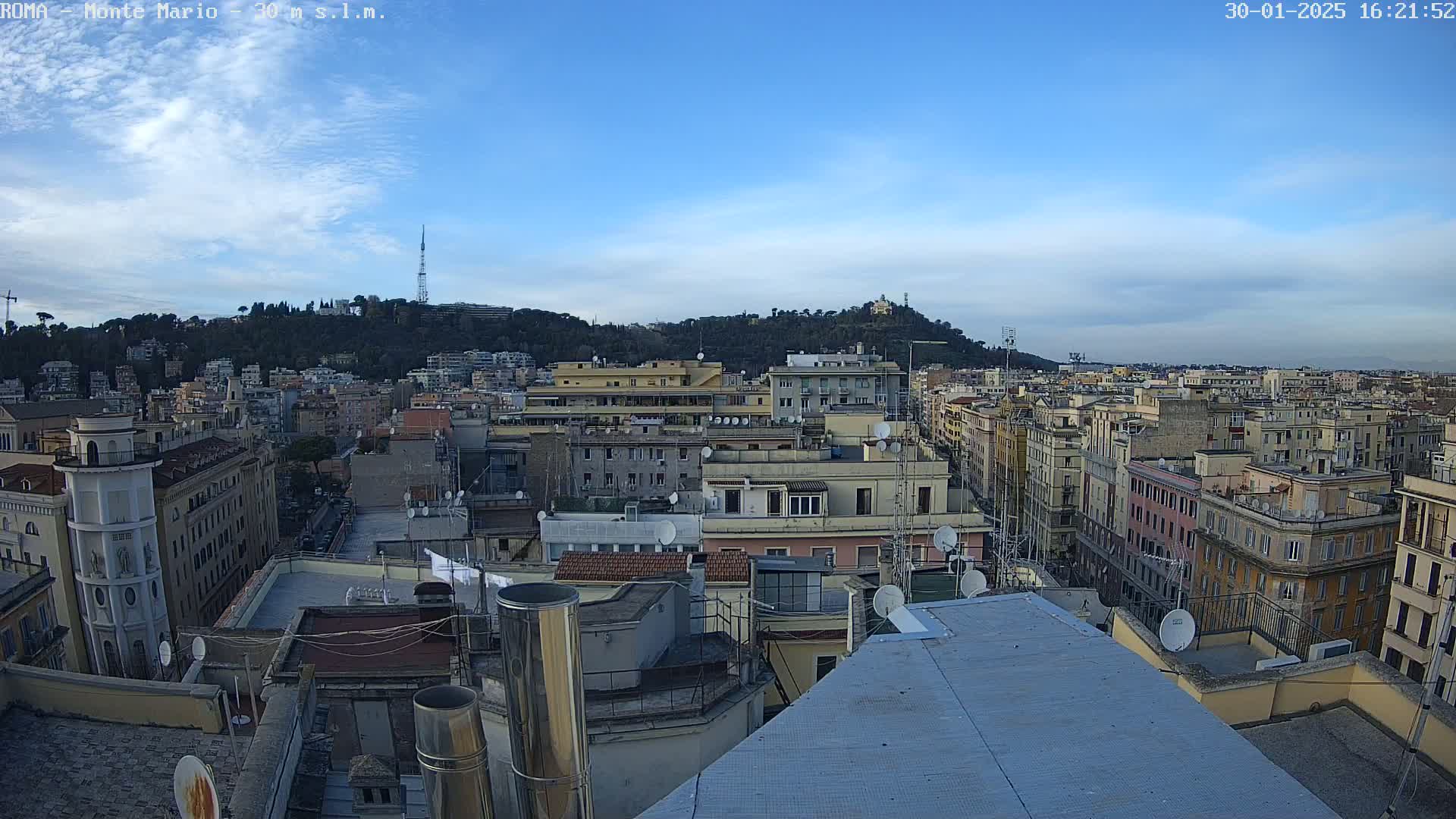 A mostly clear, daytime view of a dense cityscape with a hill in the background, showing many rooftops with satellite dishes and chimneys.