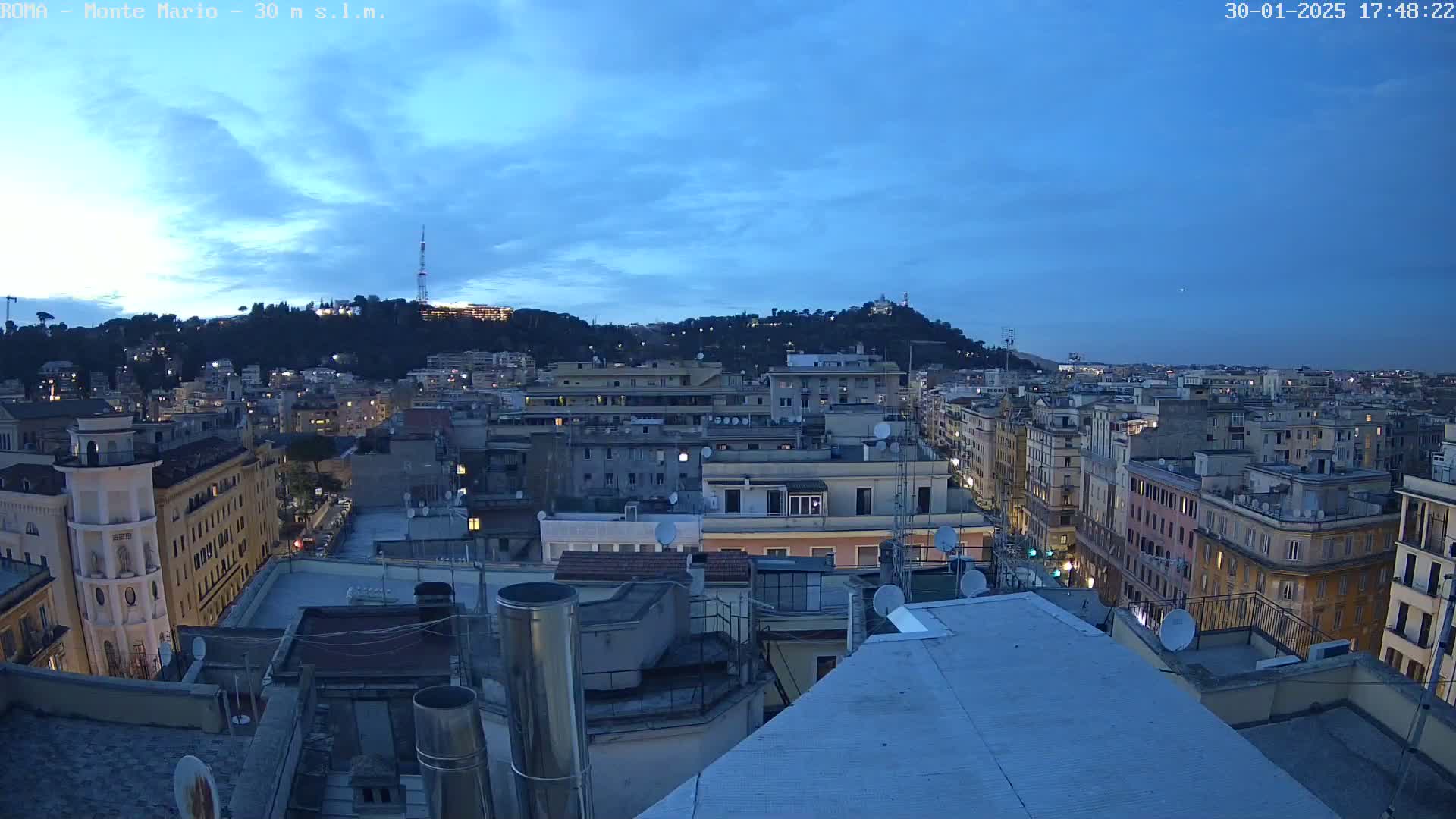 A twilight cityscape view shows many buildings stretching to a hill in the distance under a mostly clear, dark blue sky.