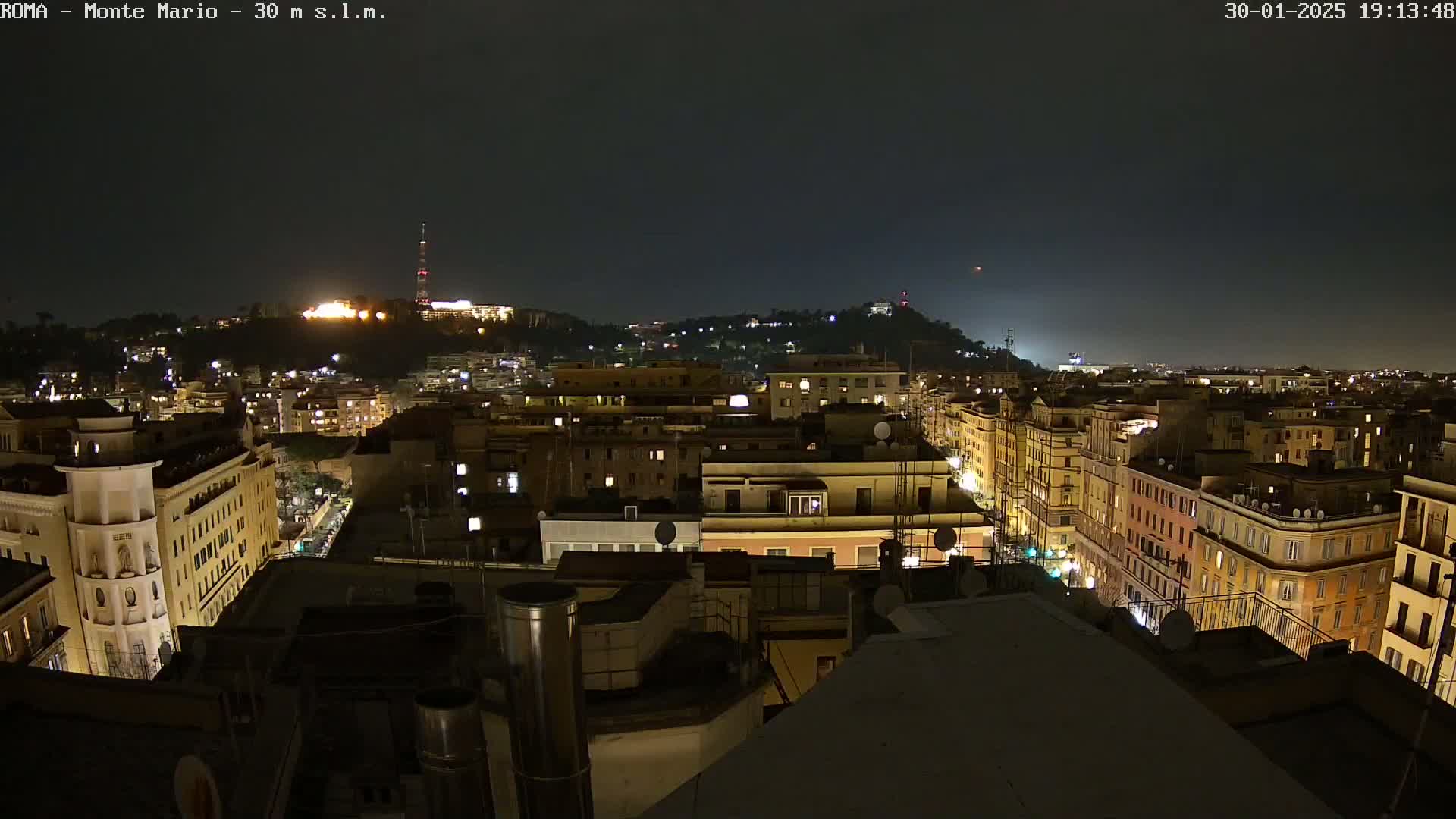 A nighttime, aerial view of a city, showing brightly lit buildings and streets, with a hilltop in the distance under a dark, clear sky.